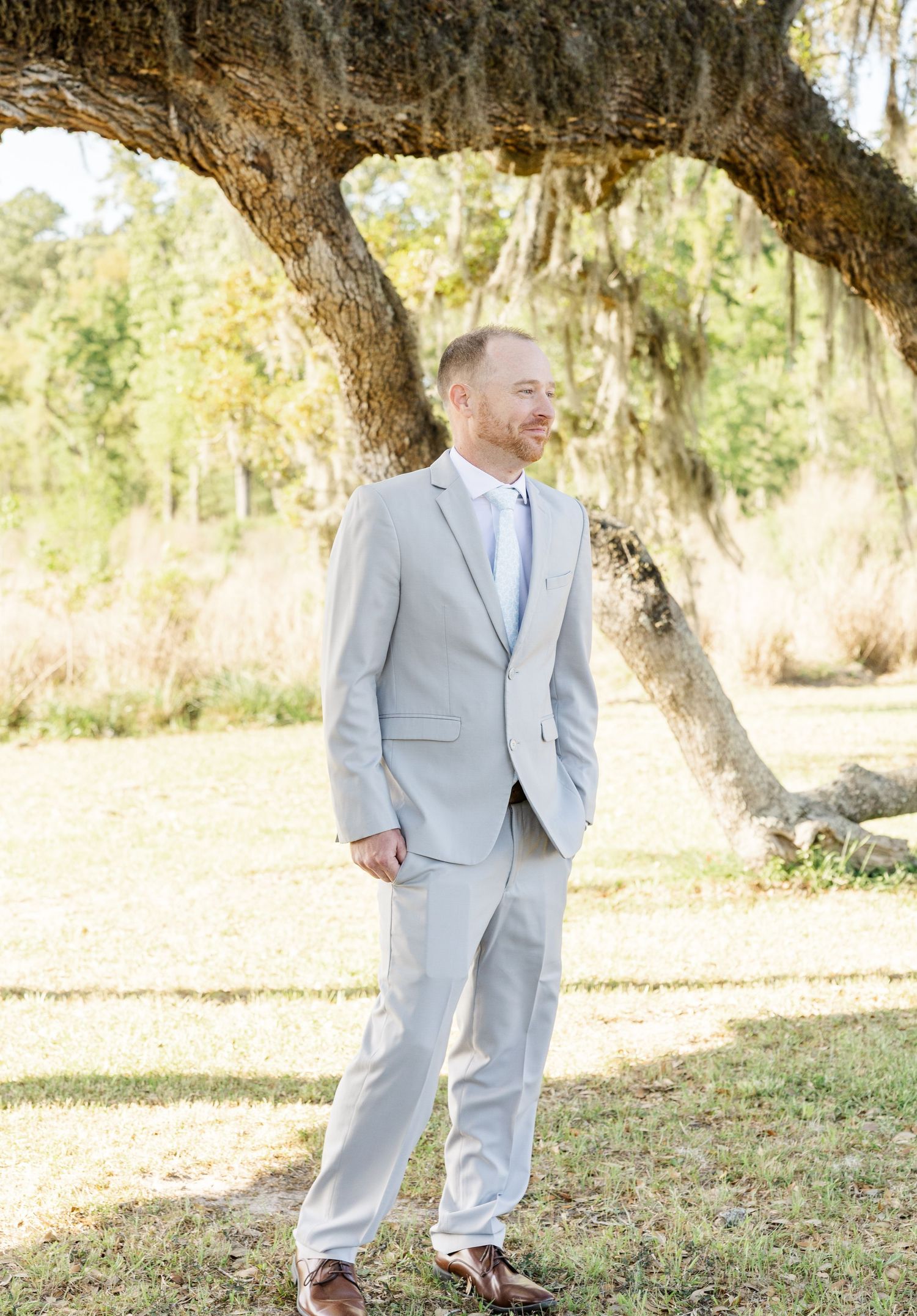 Smiling groom in light gray suit standing beneath a large Spanish moss oak tree outdoors.