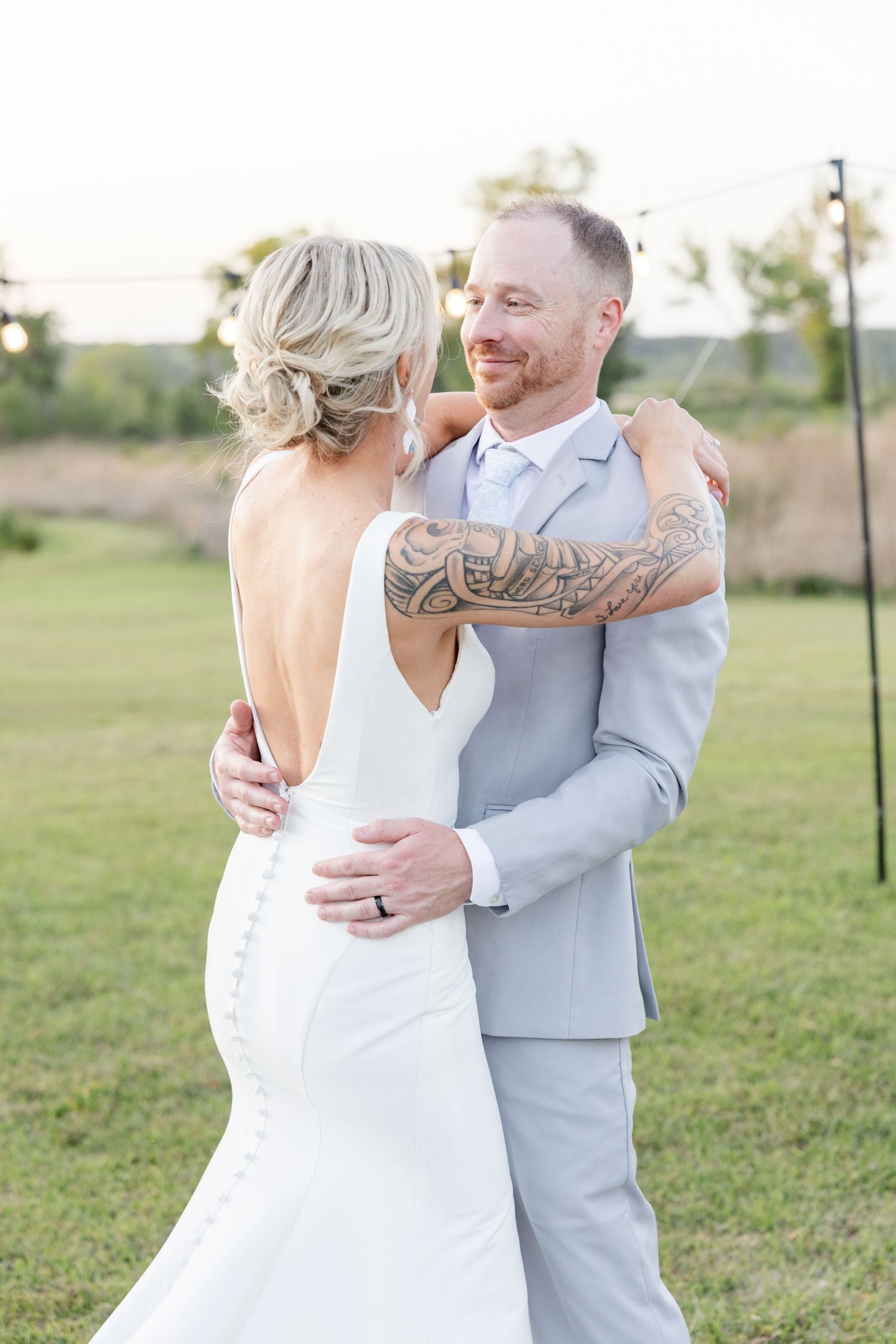 Groom with tattooed arm in gray suit embraces bride in backless white gown during outdoor first dance.