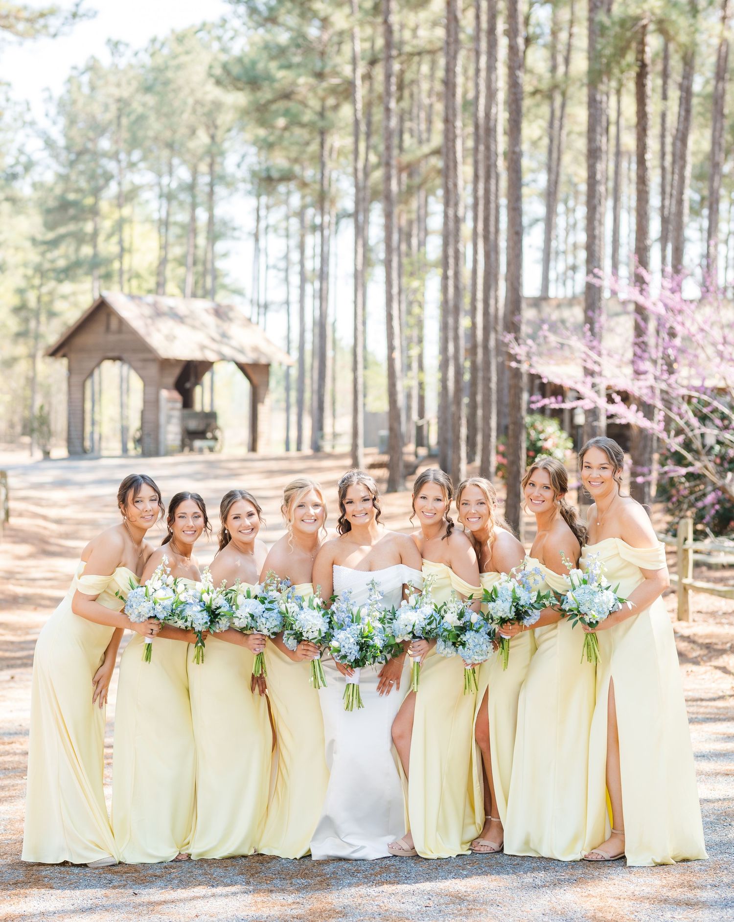 Bride stands with seven bridesmaids in pale yellow off-shoulder gowns holding blue floral bouquets.
