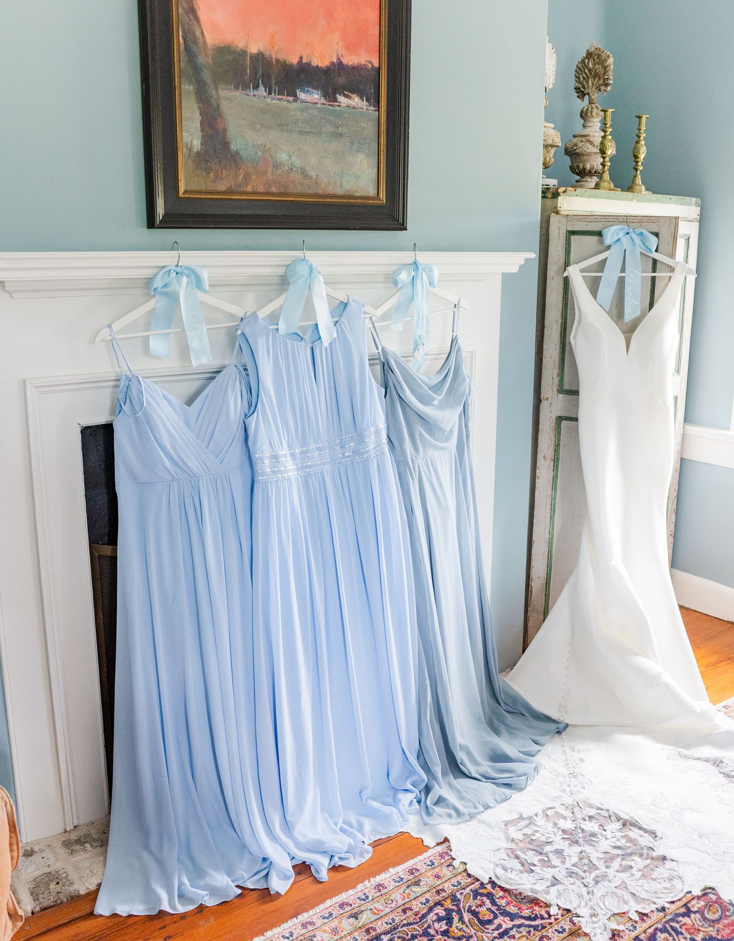 Light blue bridesmaid dresses and white wedding gown hanging near fireplace in elegant blue room.
