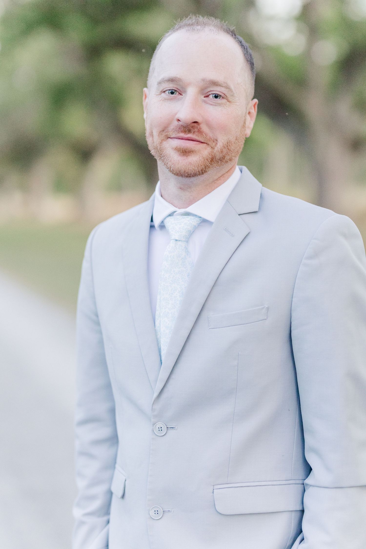 Handsome groom in a light gray suit and tie poses outdoors with a warm smile at his wedding.