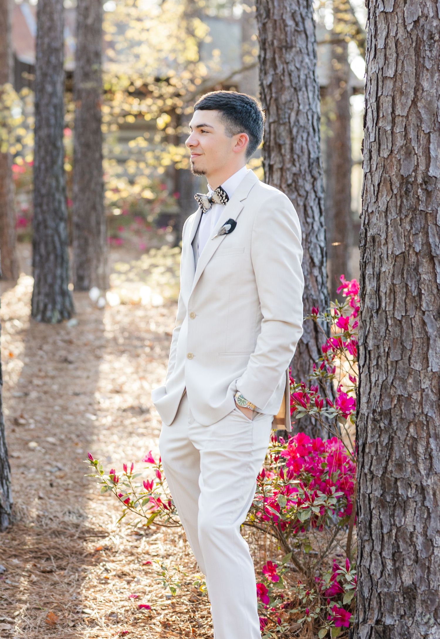 Handsome groom in cream suit with bow tie posing among pine trees with pink azaleas at outdoor wedding.