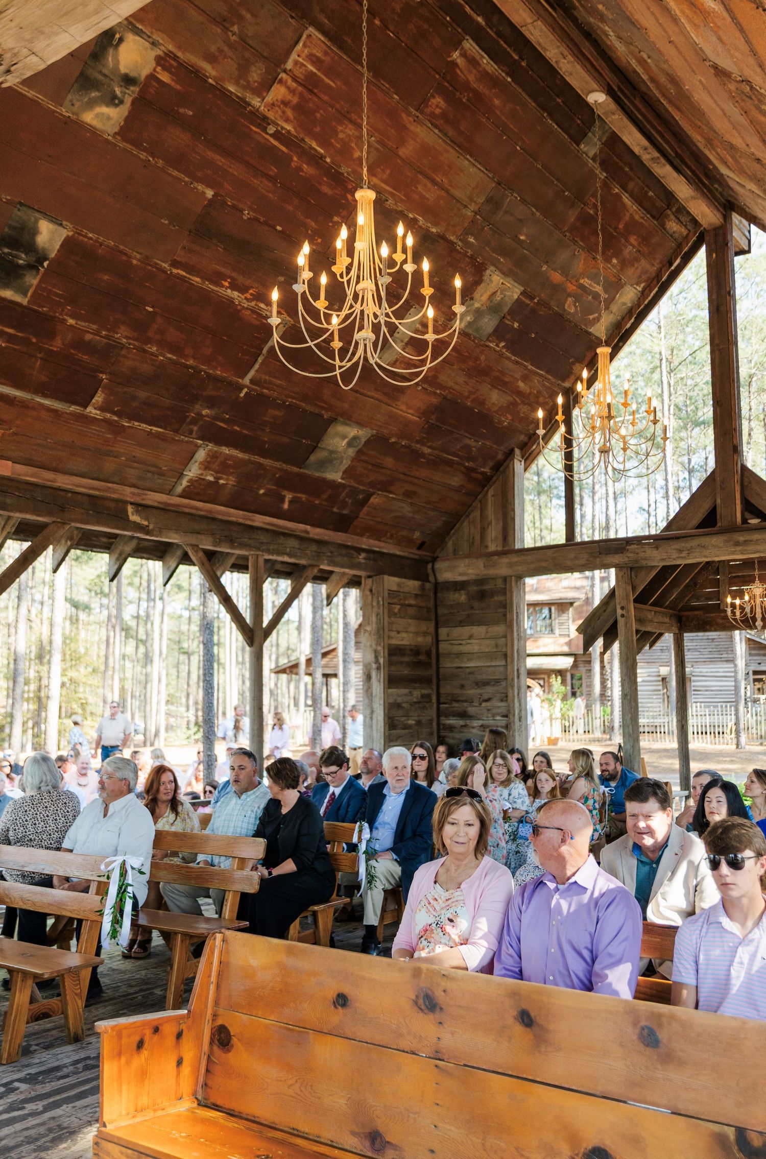 Wedding guests seated on wooden pews under rustic barn chapel with elegant chandelier lighting.