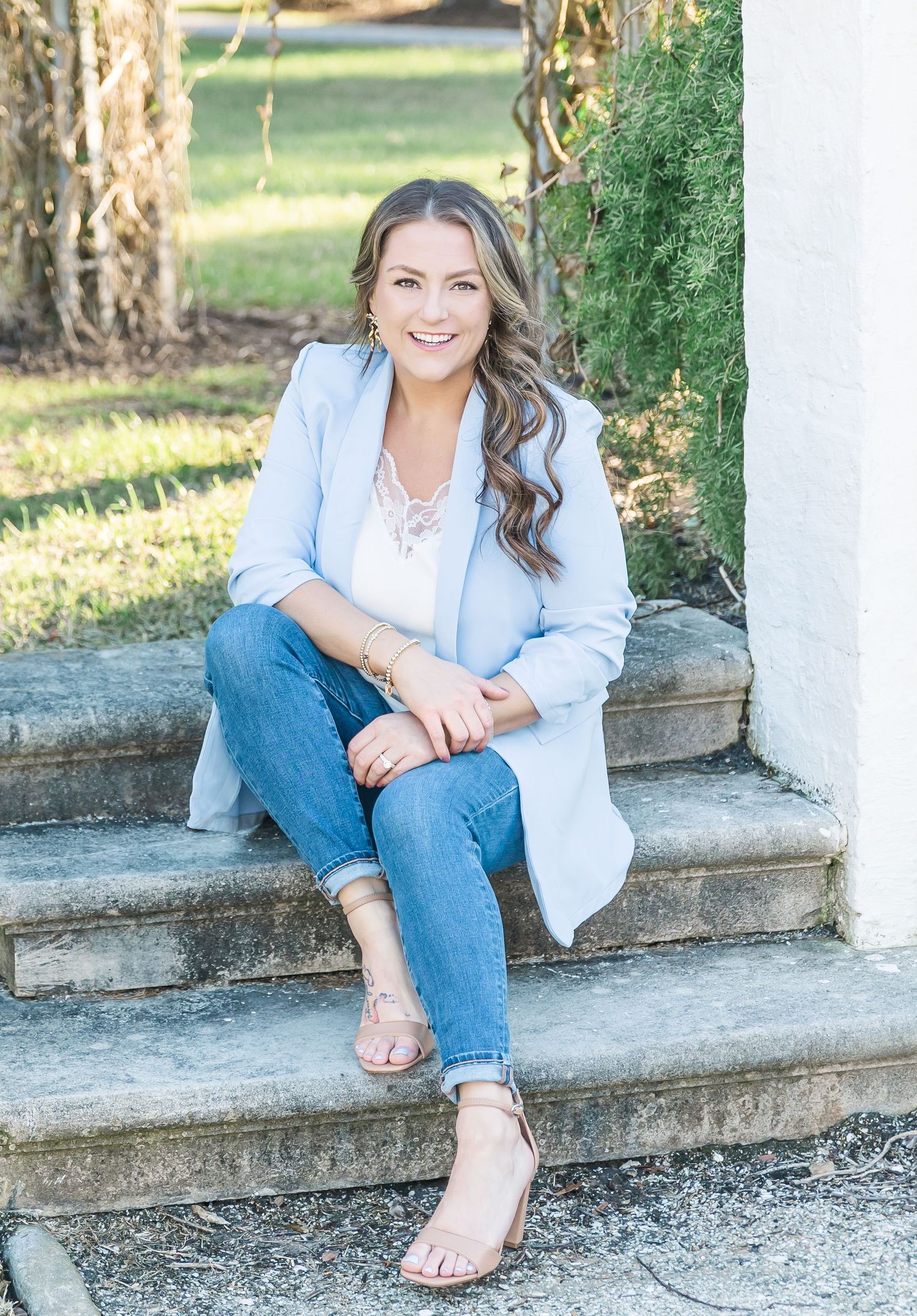 Person in light blue blazer and jeans sits casually on outdoor stone steps in a natural garden setting.