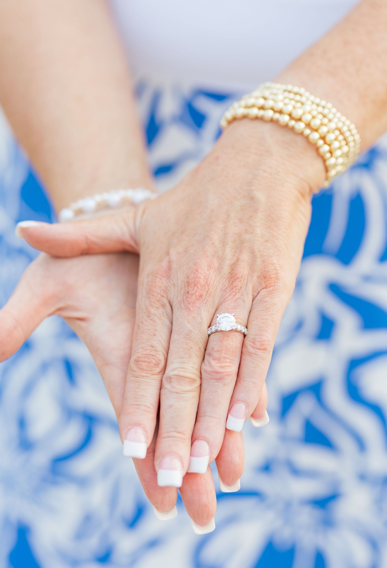 A close-up of a hand wearing a pearl bracelet and engagement ring against a blue floral pattern.