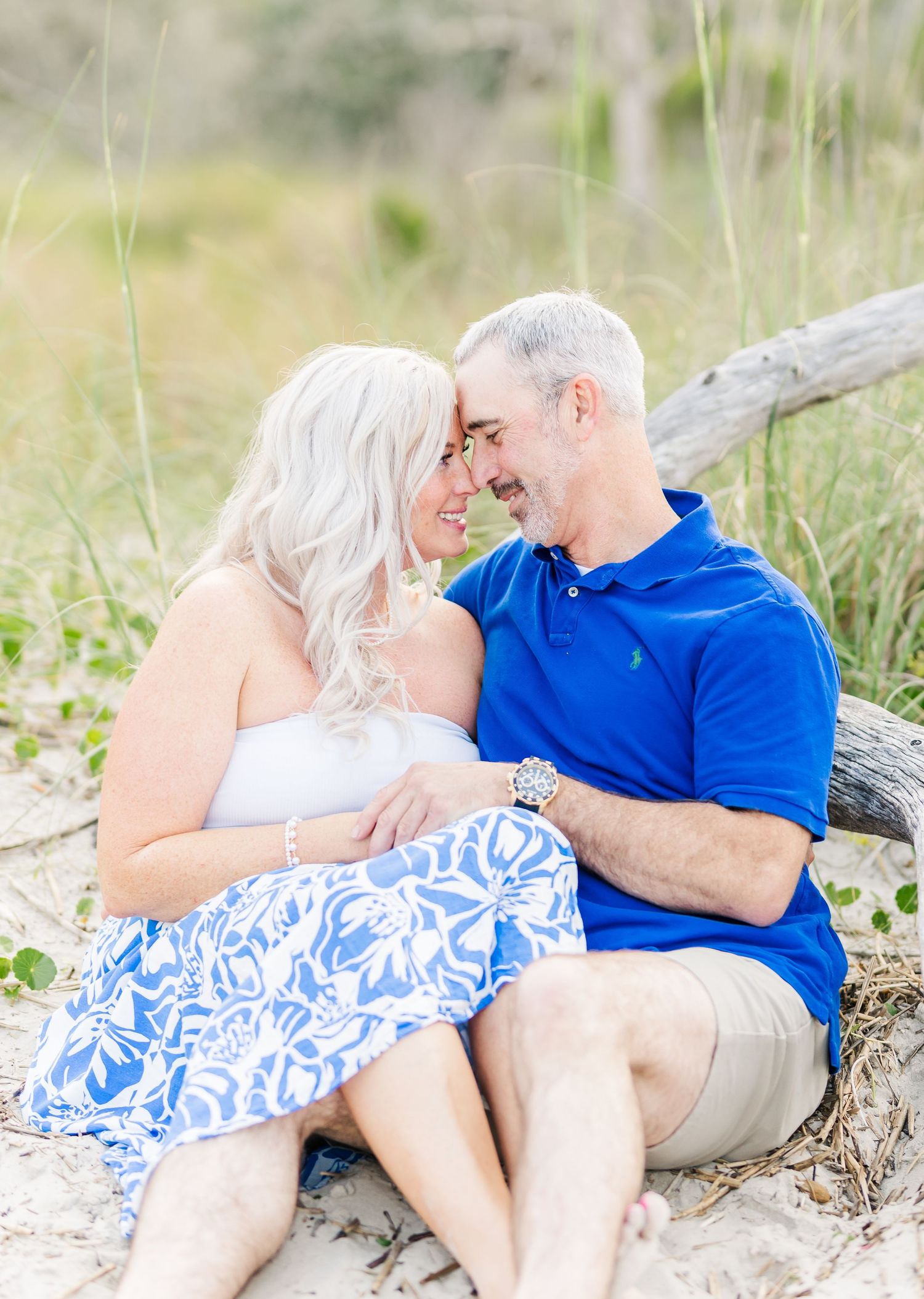 A couple in coordinated blue and white outfits share an intimate moment while sitting on driftwood at the beach.