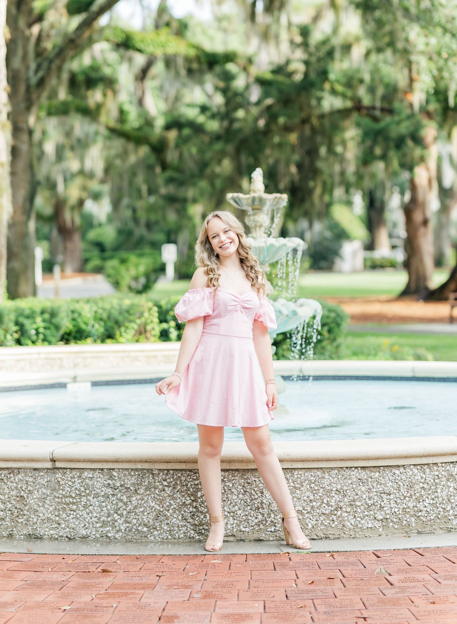A person in a pink off-shoulder dress poses by an ornate stone fountain in a lush garden setting.
