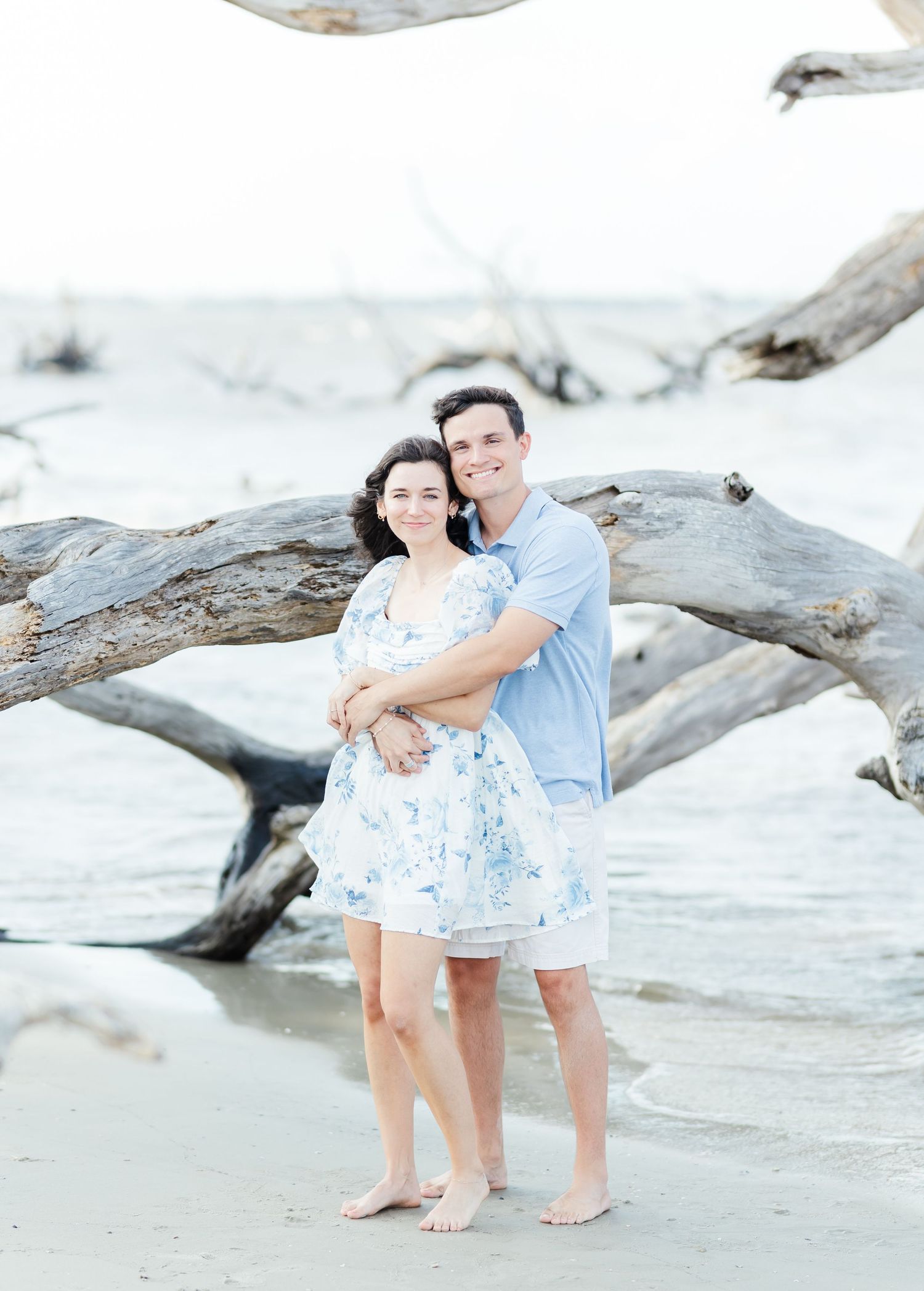 Couple in coordinating blue and white outfits embracing on a driftwood-covered beach.
