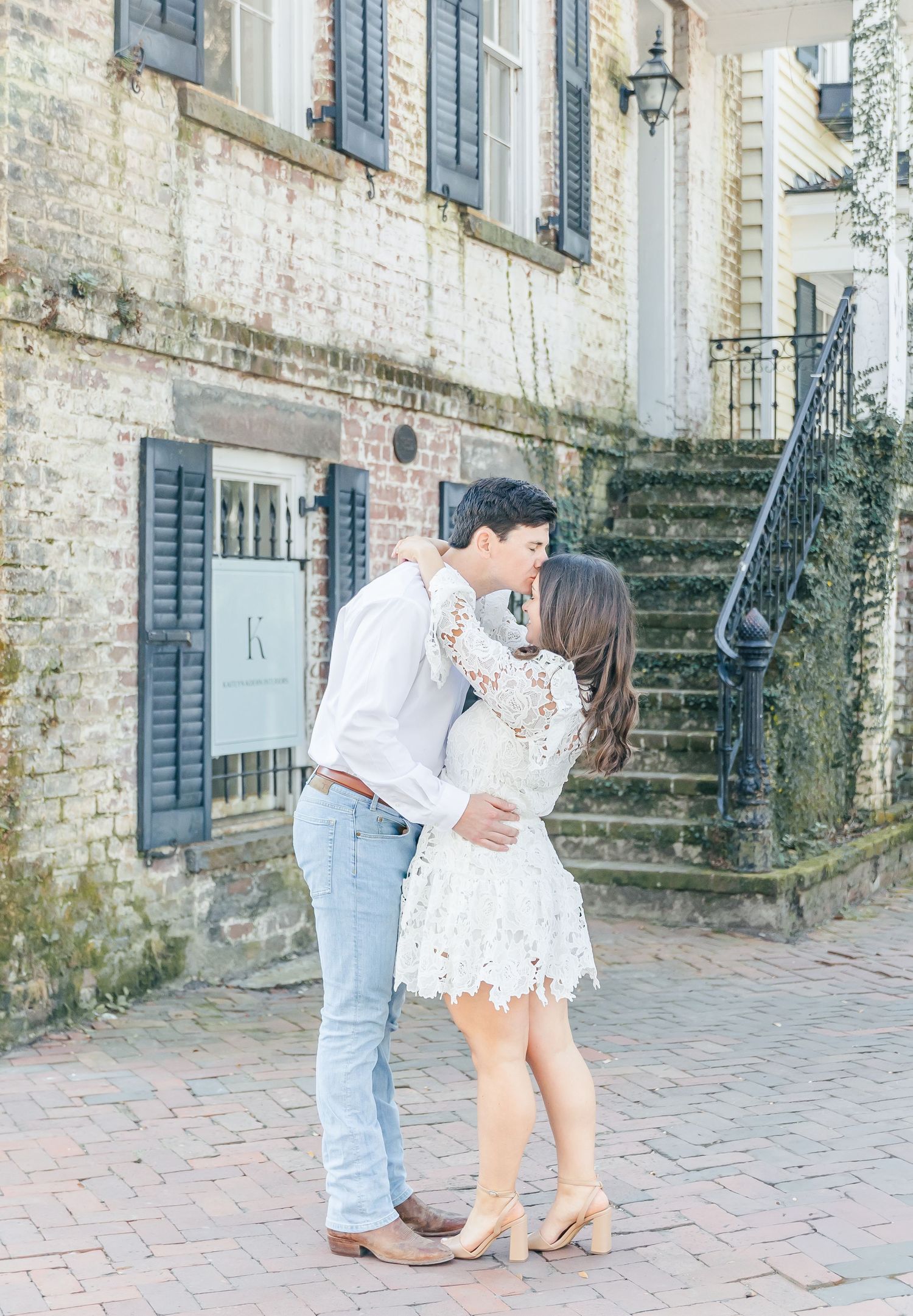 A couple shares a tender moment on brick steps outside a historic building with blue shuttered windows.