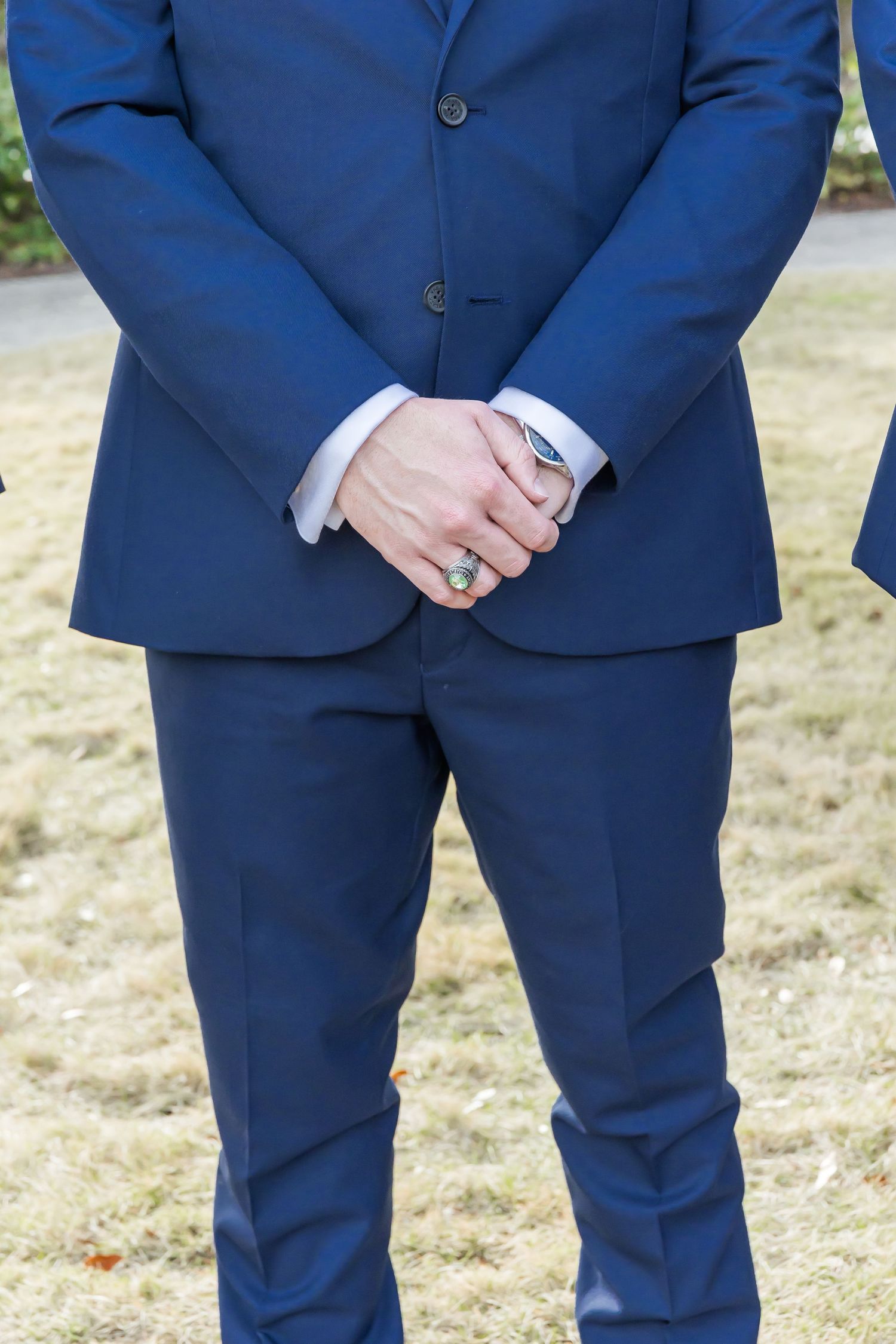 Close-up detail of a groom in a navy blue suit with clasped hands showing wedding ring details.