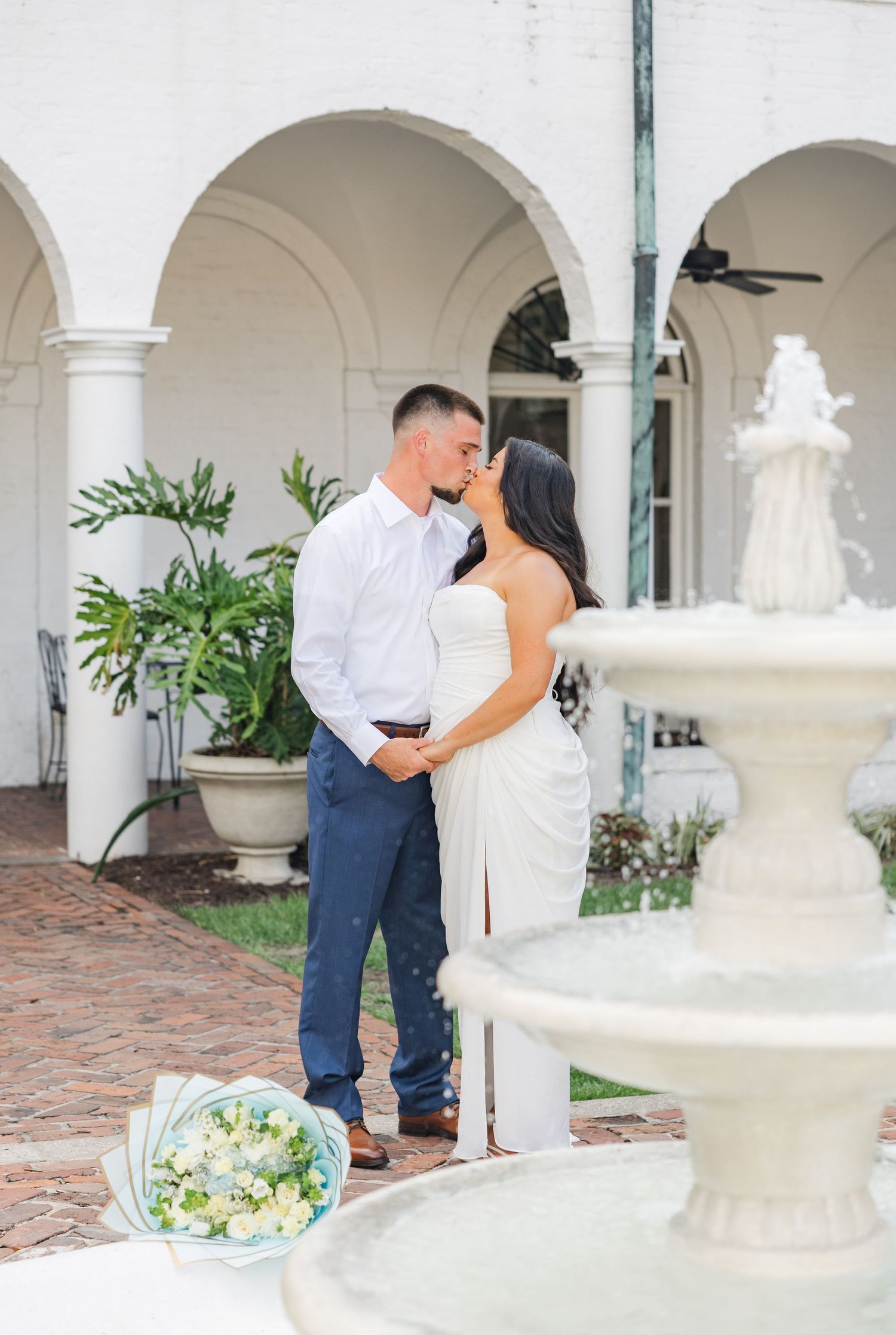 Newly engaged couple shares a kiss near ornate white fountain with flower bouquet nearby.