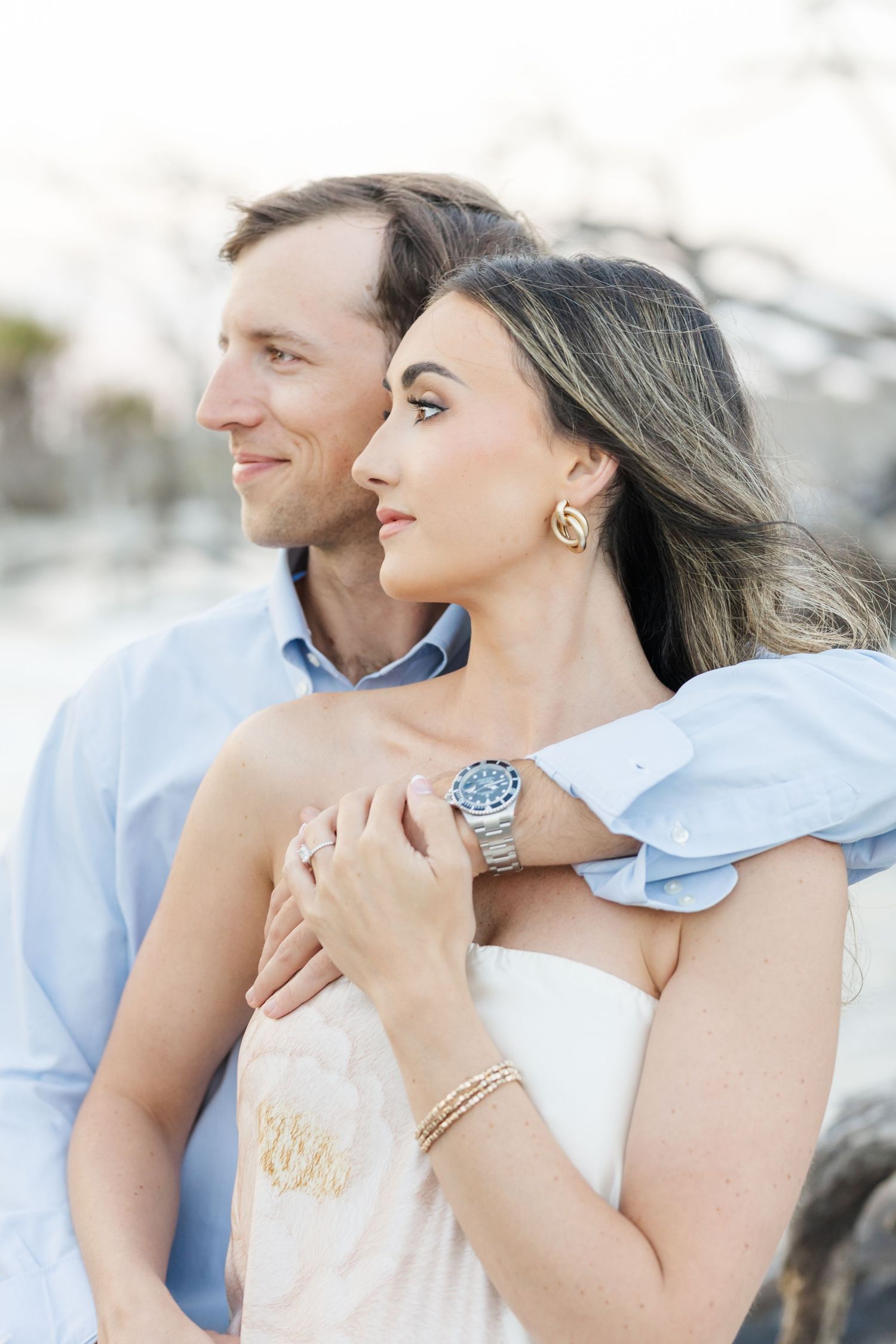 Couple embracing outdoors, woman in strapless dress wearing a Rolex watch, man in blue shirt holding her close.