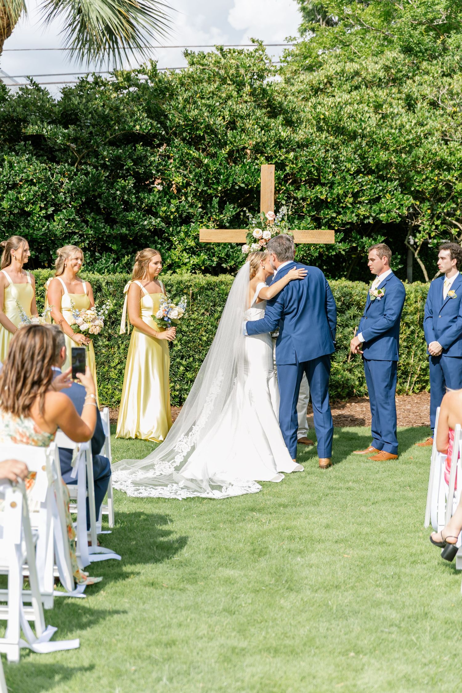 Wedding party arranged on grassy lawn with palm trees, featuring navy suits and pale yellow dresses.