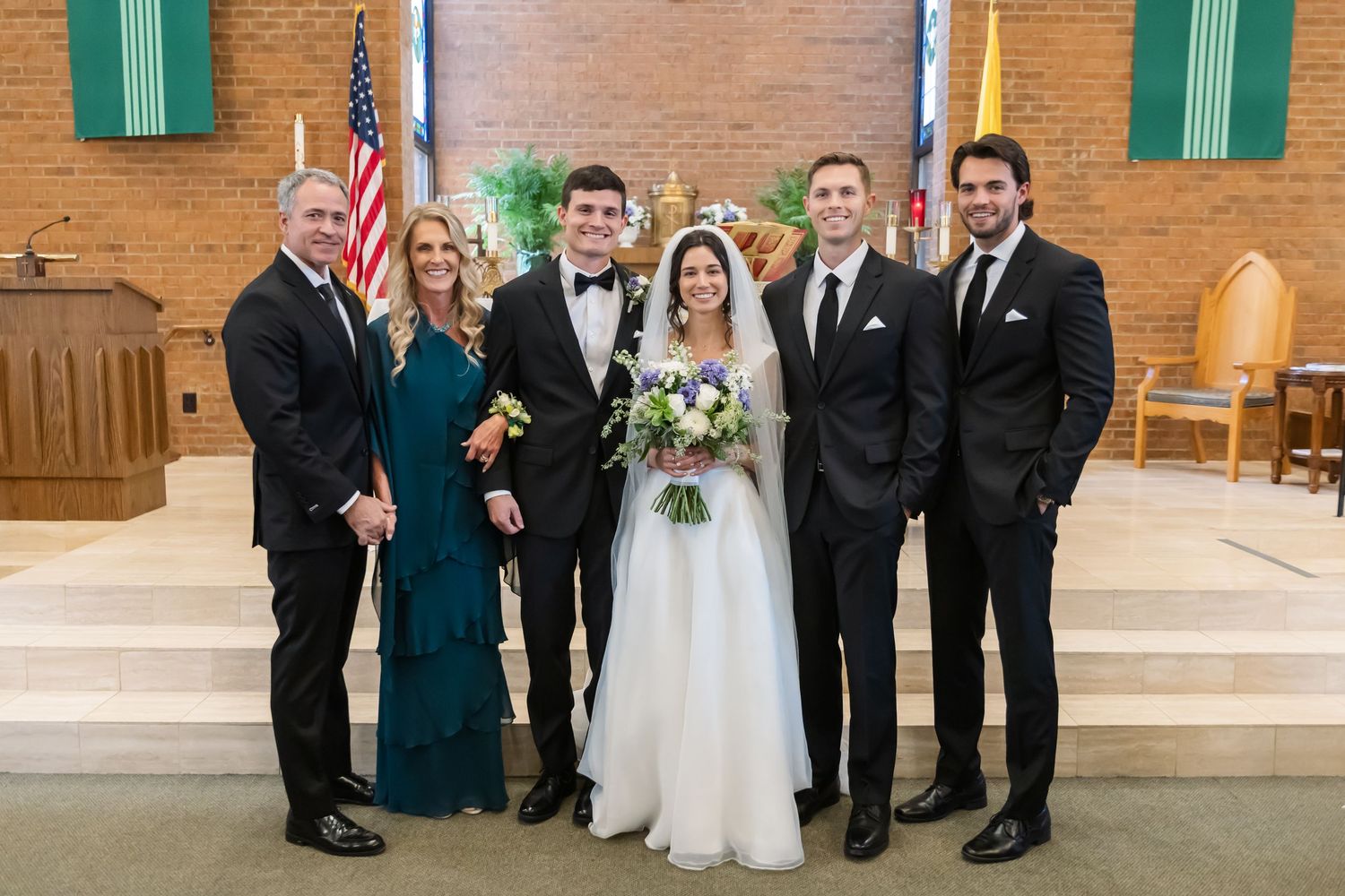 Wedding party members in formal attire pose together inside a brick church with green shutters and American flag.