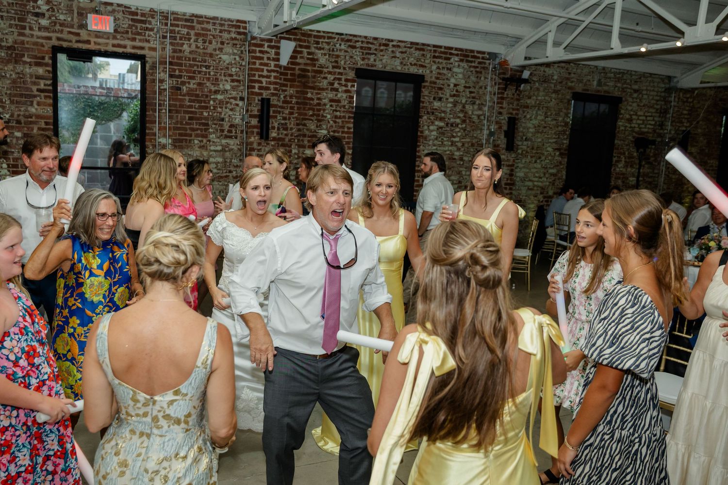 Wedding guests dance and celebrate on the dance floor at an indoor reception with exposed brick walls and white ceiling.