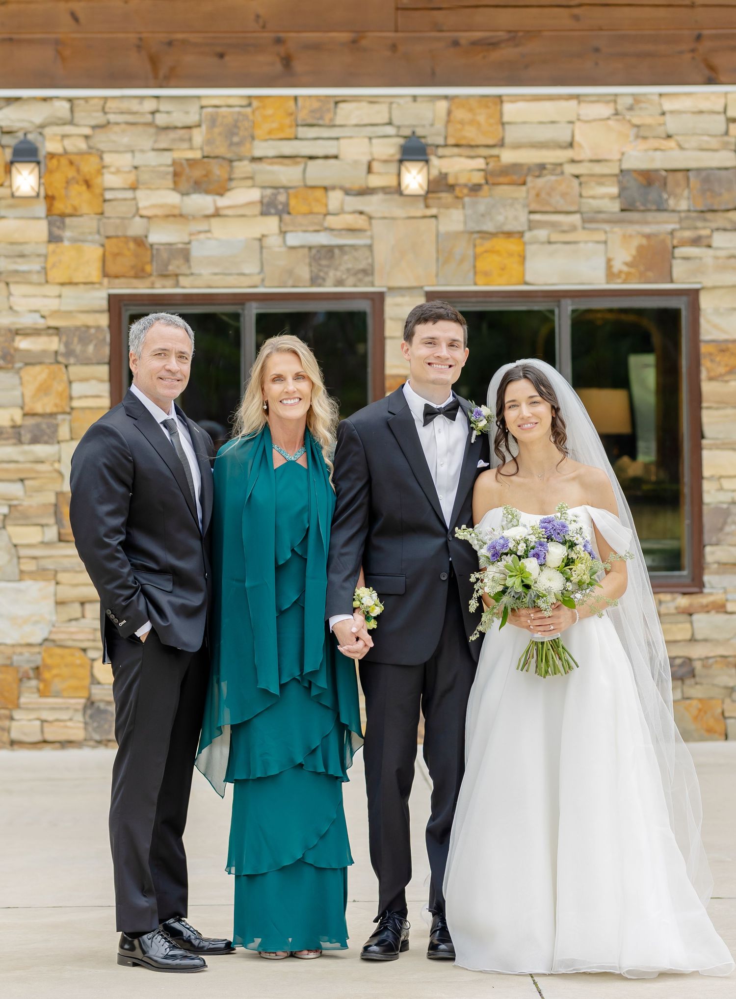 Wedding party poses outdoors against stone wall backdrop in formal attire with green and black color scheme.