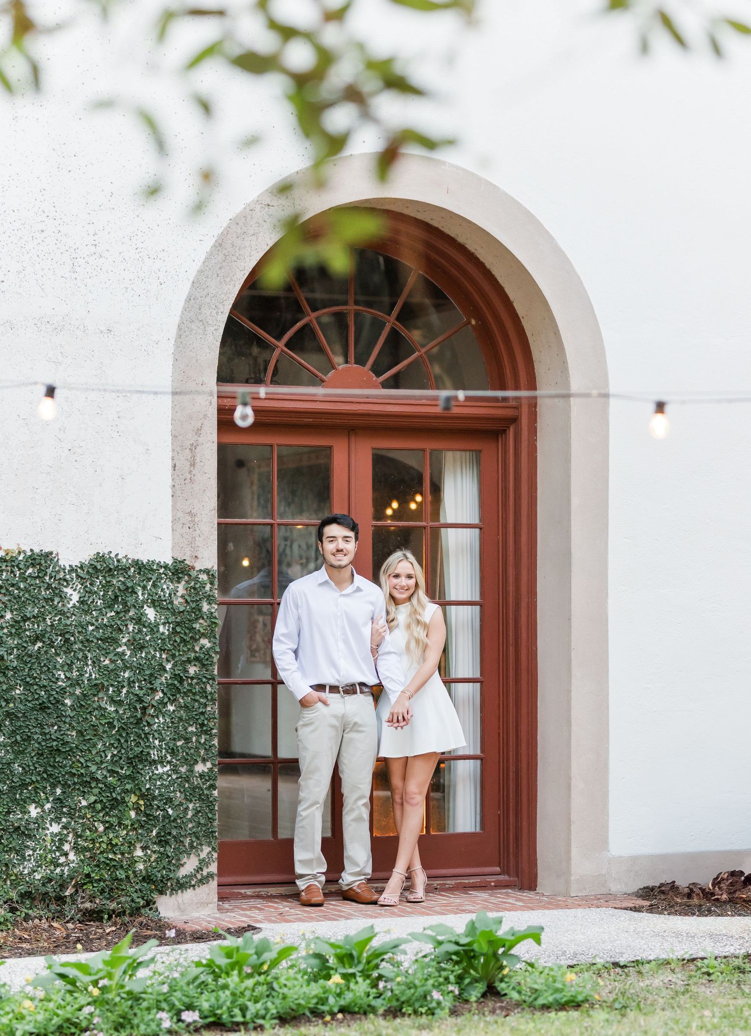 A couple stands together in a Spanish-style arched doorway with string lights overhead.