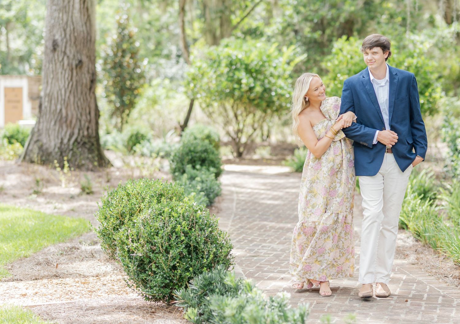 A couple strolls together on a garden path surrounded by manicured shrubs and trees in soft natural light.