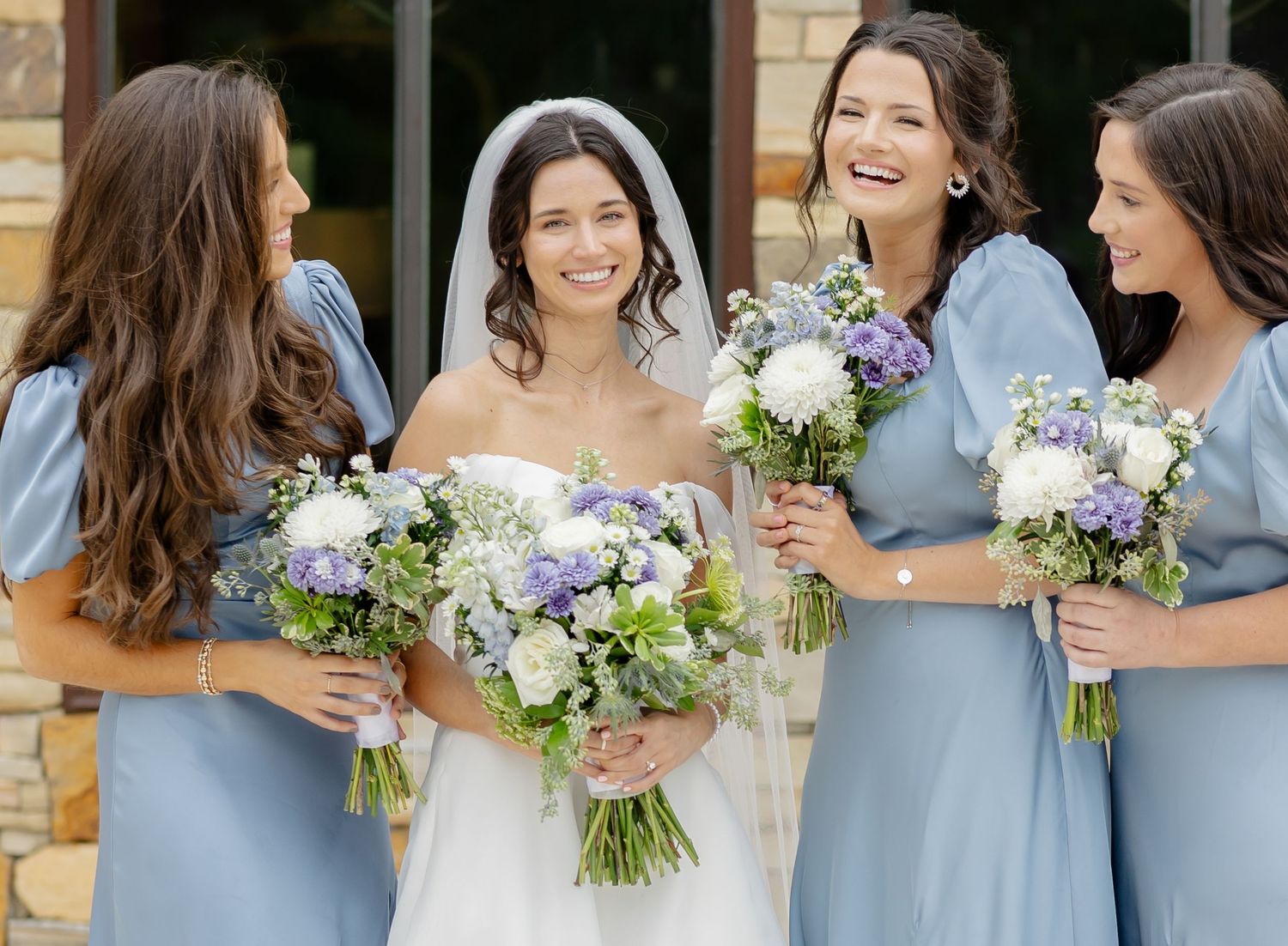 Bridesmaids in light blue dresses stand together holding white and purple floral bouquets at an outdoor wedding.