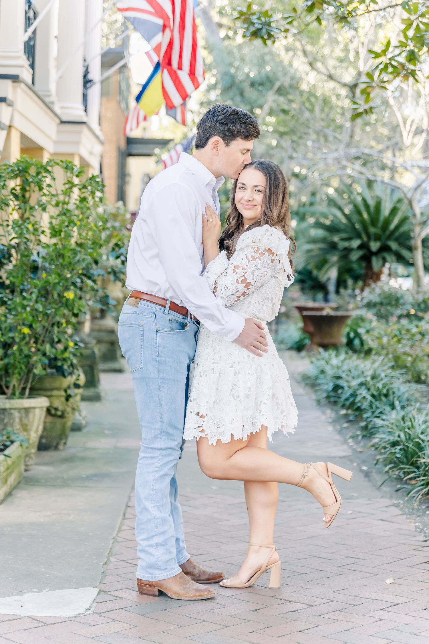 Playful engagement photo with woman in lace dress lifting her foot while embracing partner.