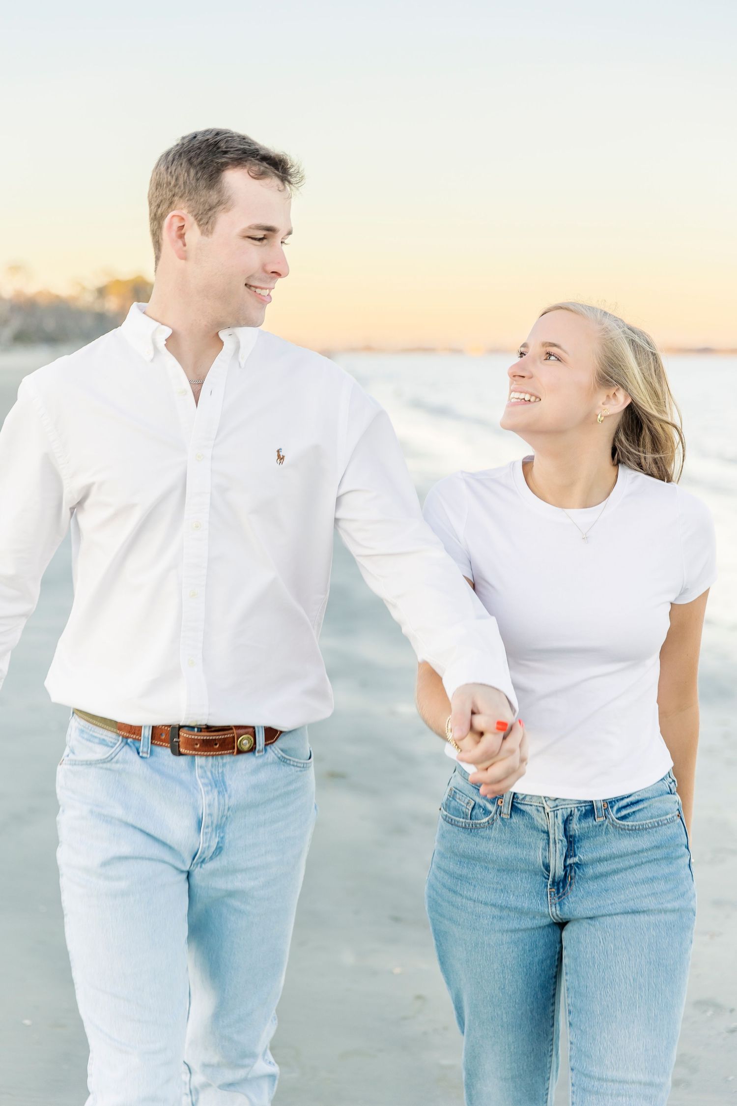 Couple in casual white and denim outfits walking together on a beach during sunset.