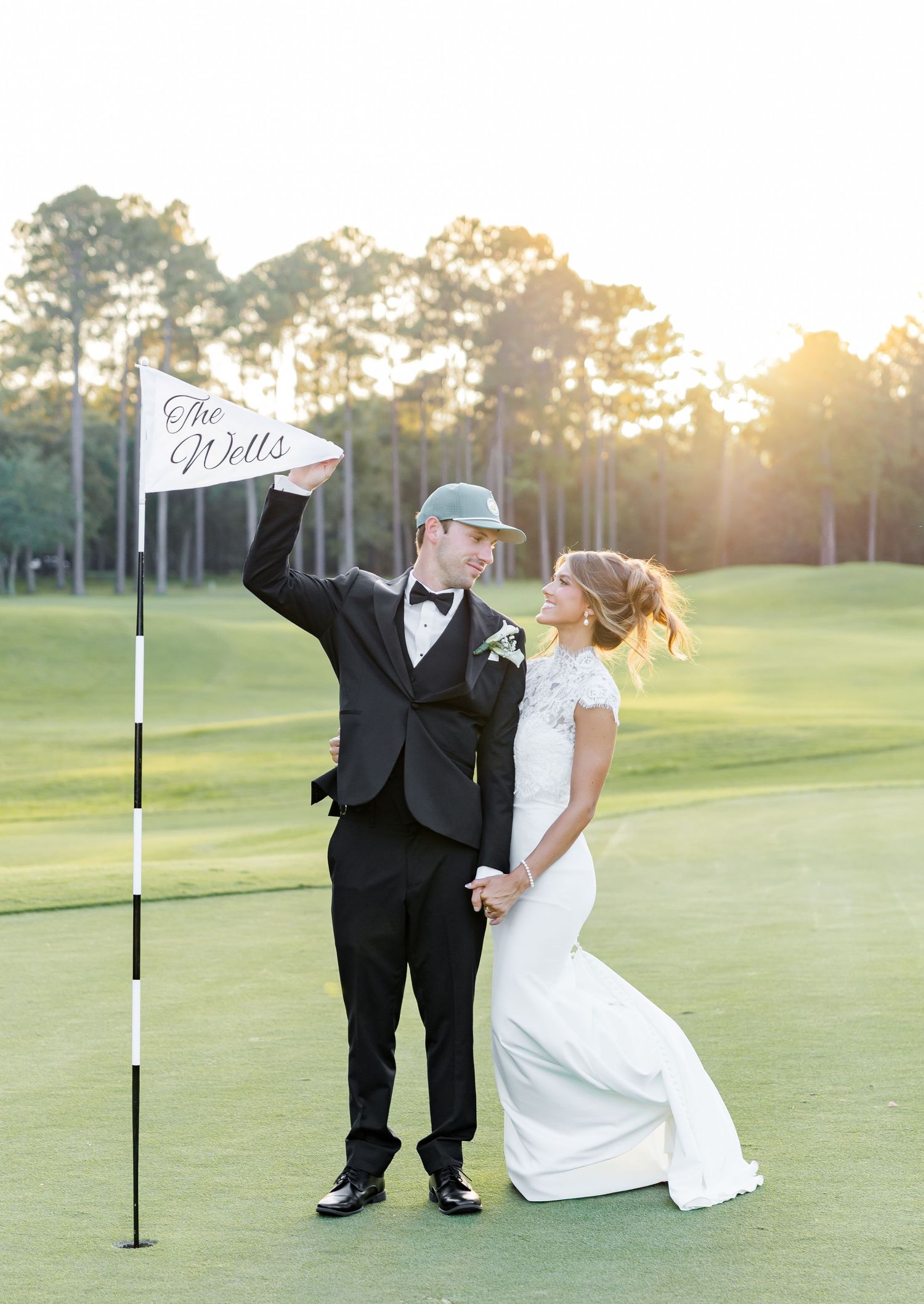 Newlywed couple posing on golf course green at sunset, groom holding flag reading The Wells, bride in white gown.