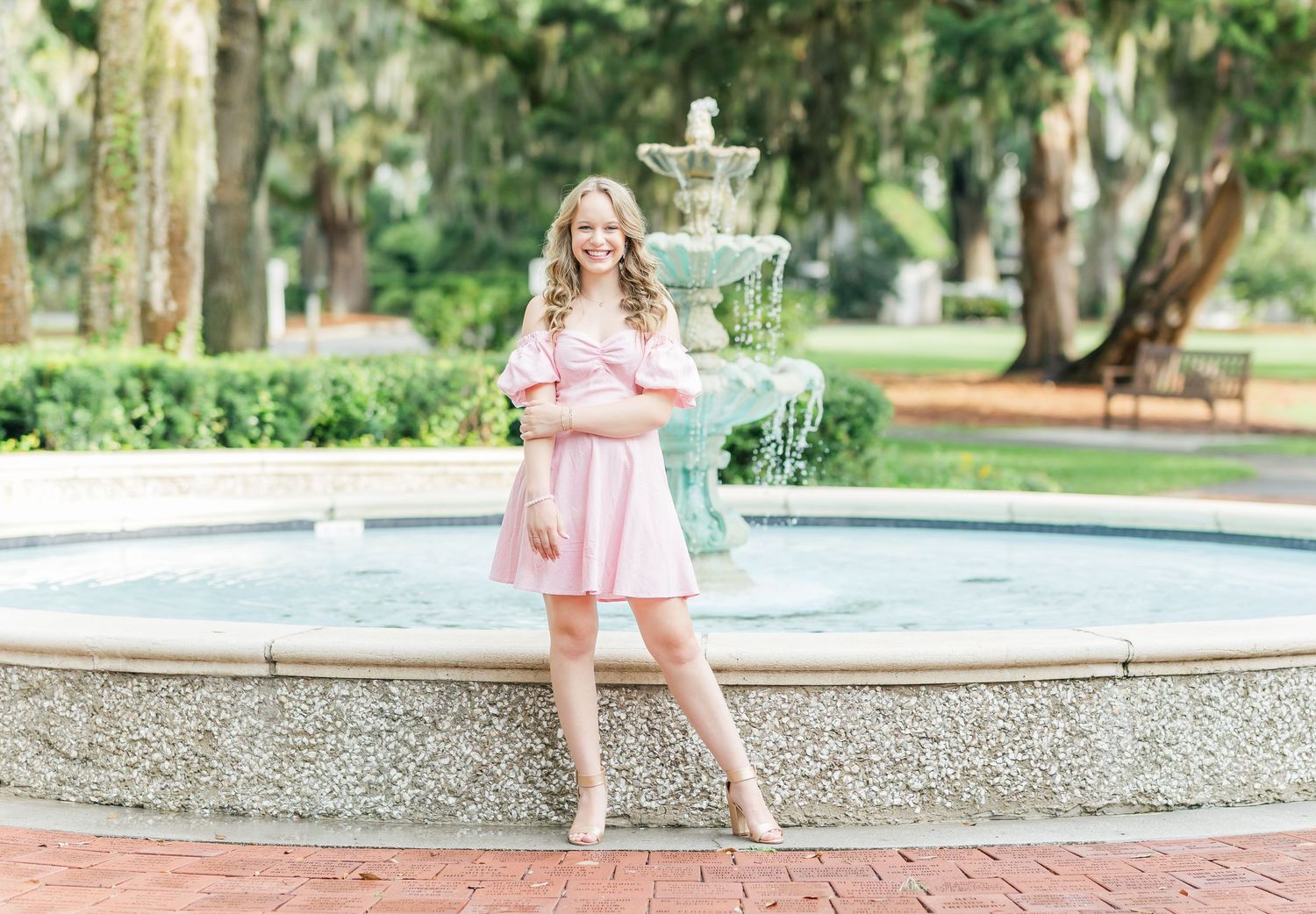 A person in a pink dress and sun hat stands gracefully in a garden setting with brick pathways and greenery.