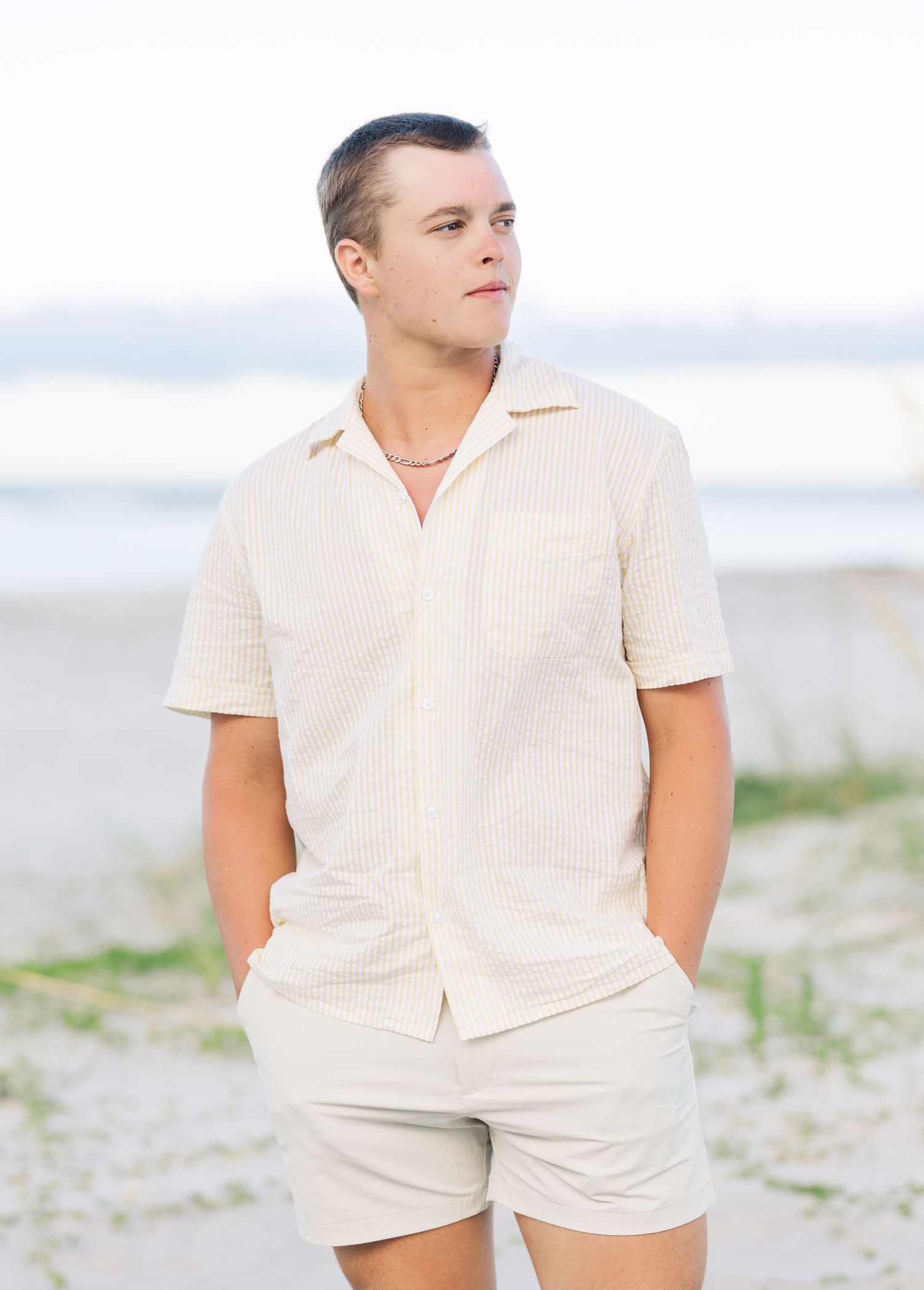Person wearing a cream colored outfit poses casually on a sandy beach in soft lighting.
