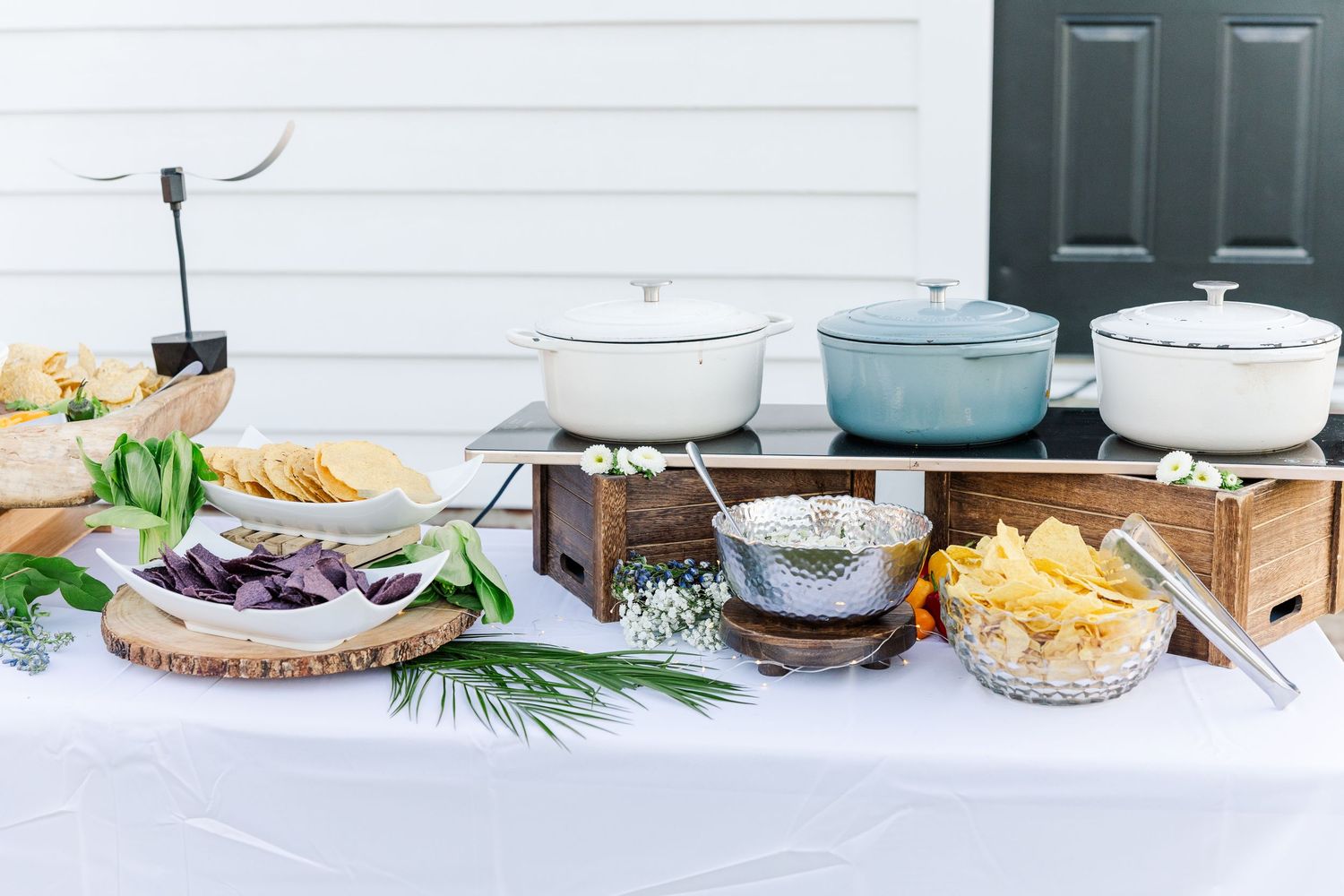 Outdoor wedding buffet table with Dutch ovens, chips, and colorful appetizers on a white tablecloth.
