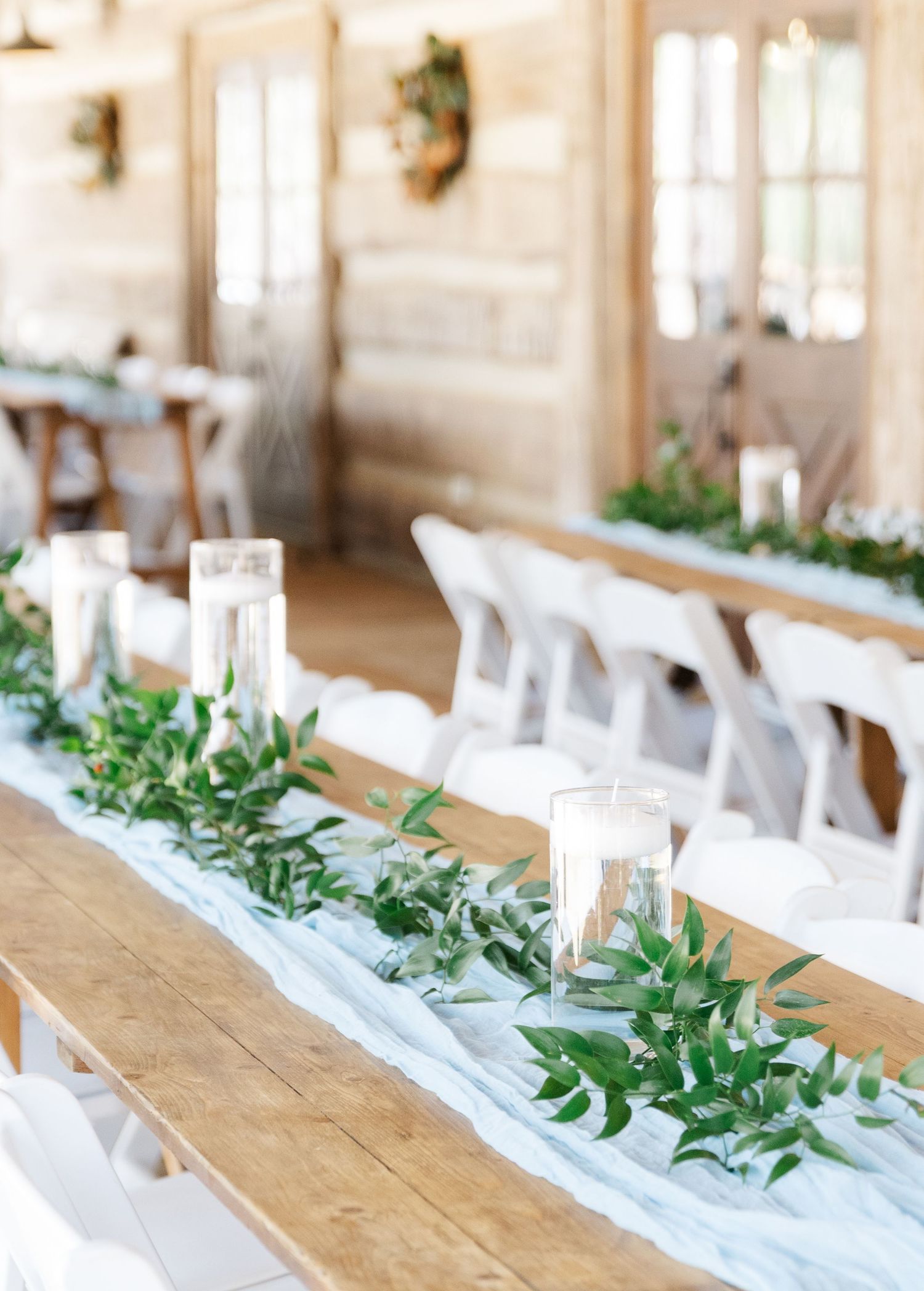 Elegant rustic barn wedding reception tables with greenery garland, white candles, and white chairs.