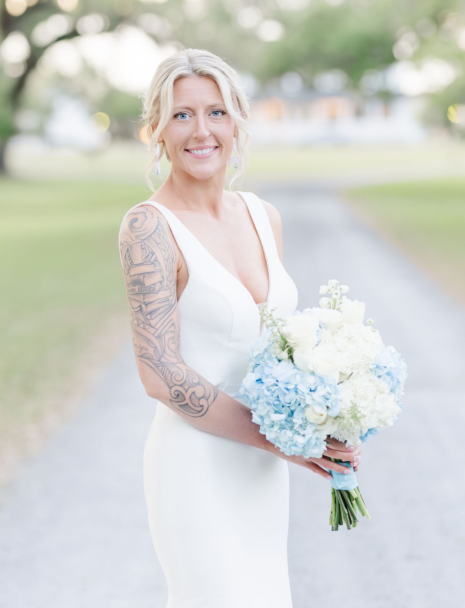 Smiling bride in white gown holding blue hydrangea bouquet with sleeve tattoo.
