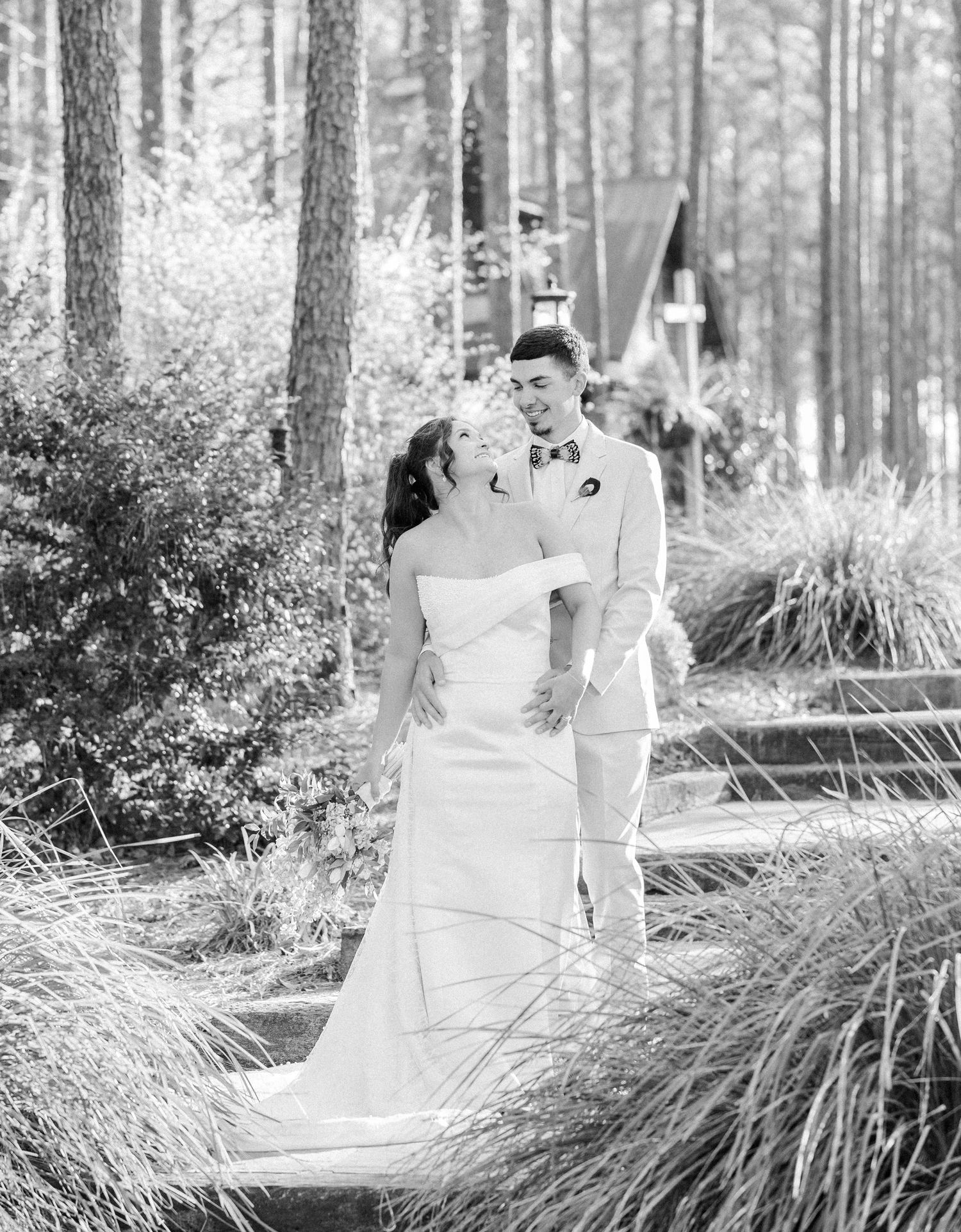 Black and white portrait of bride and groom laughing together near pine trees and a small chapel at wedding venue.