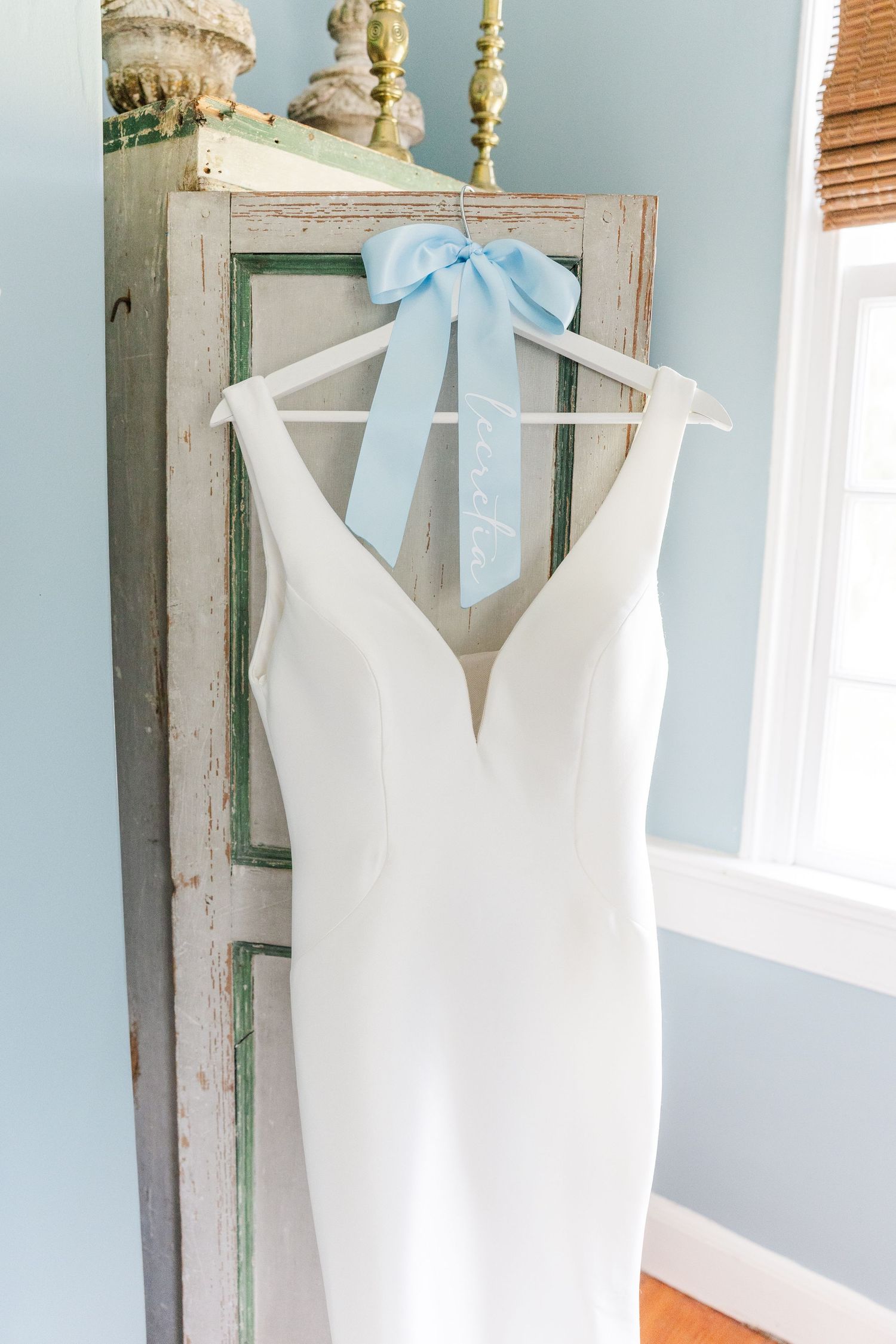 White wedding gown with deep V-neck hanging on hanger with light blue bow ribbon against rustic green cabinet.