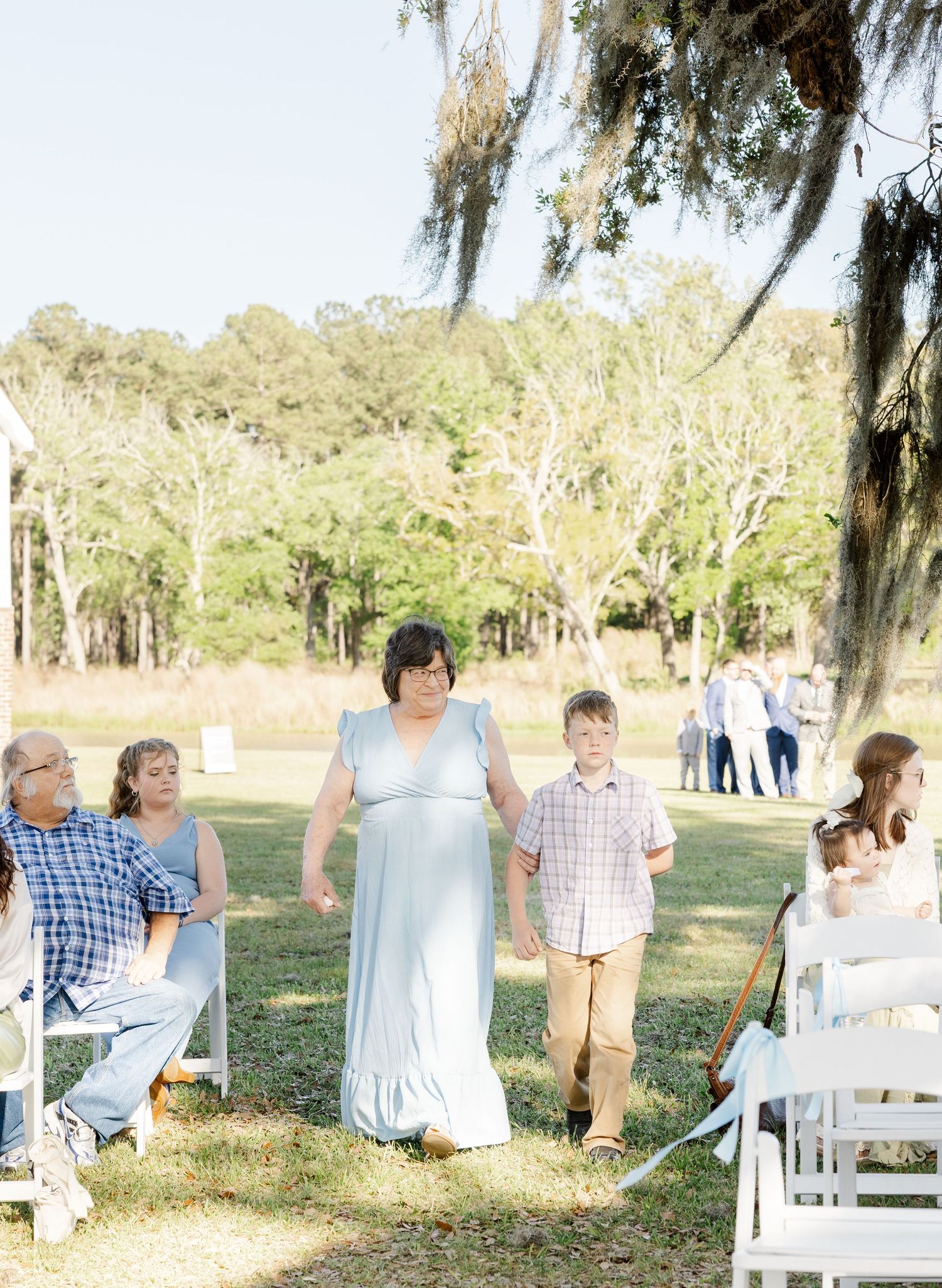 Woman in light blue dress walking down outdoor wedding aisle with young boy under Spanish moss oak trees.
