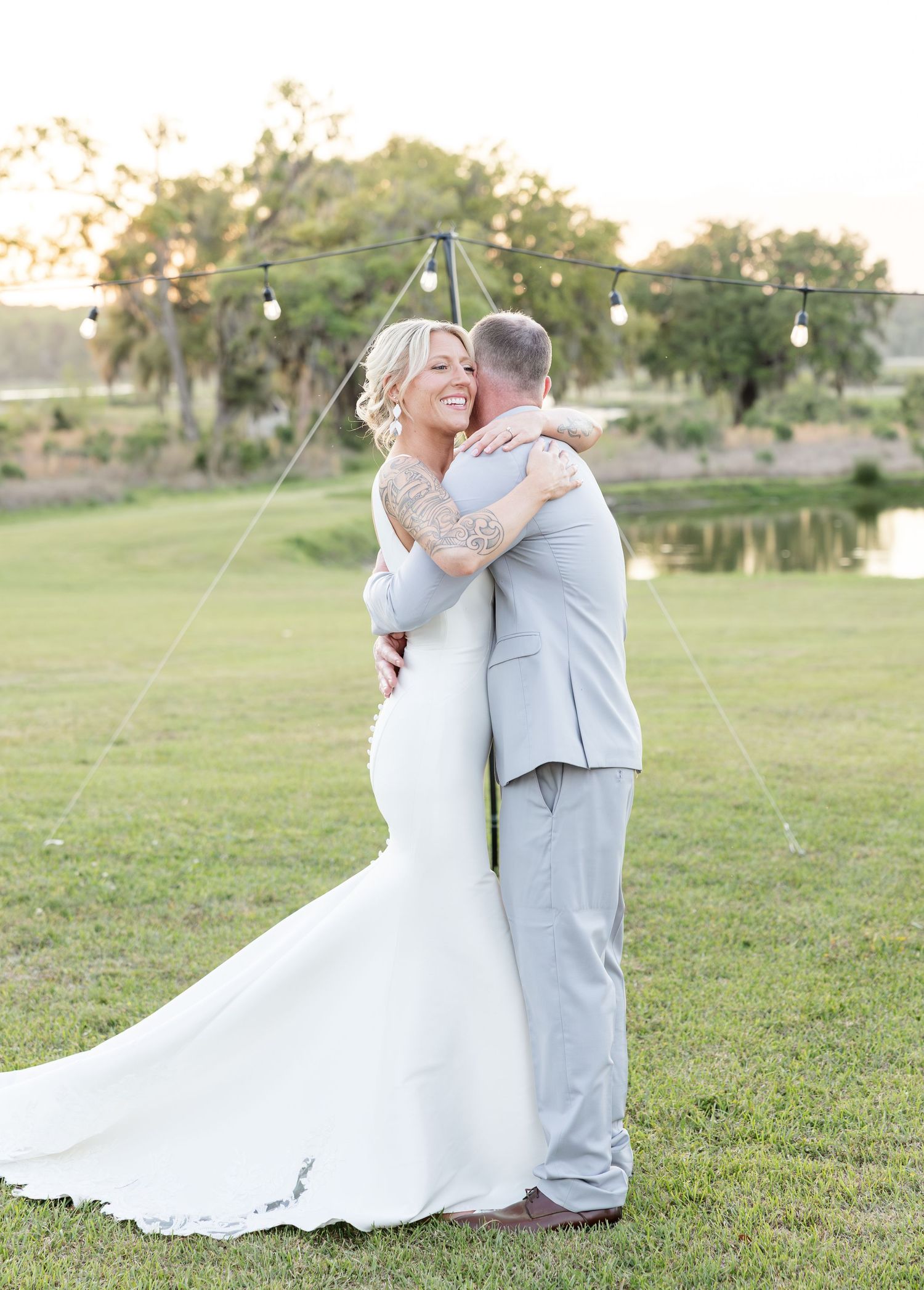 Newlywed couple embracing and smiling on green lawn with string lights and pond at golden hour.