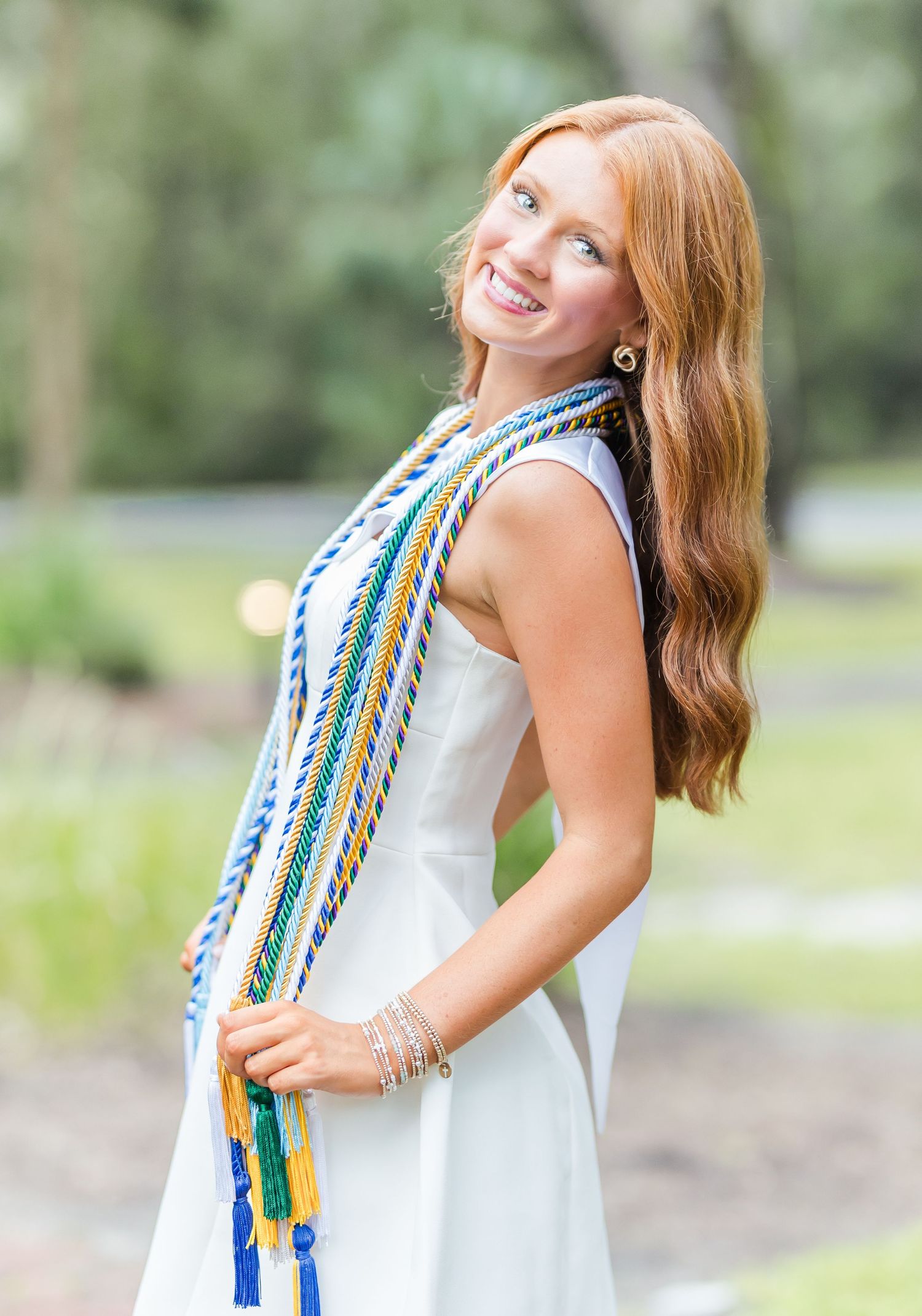 Graduate wearing a white dress and colorful graduation stole smiles outdoors during a portrait session.
