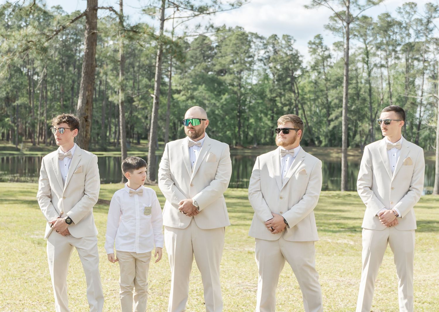 Groomsmen and young boy in light beige suits stand in line on grassy lawn with pine trees.