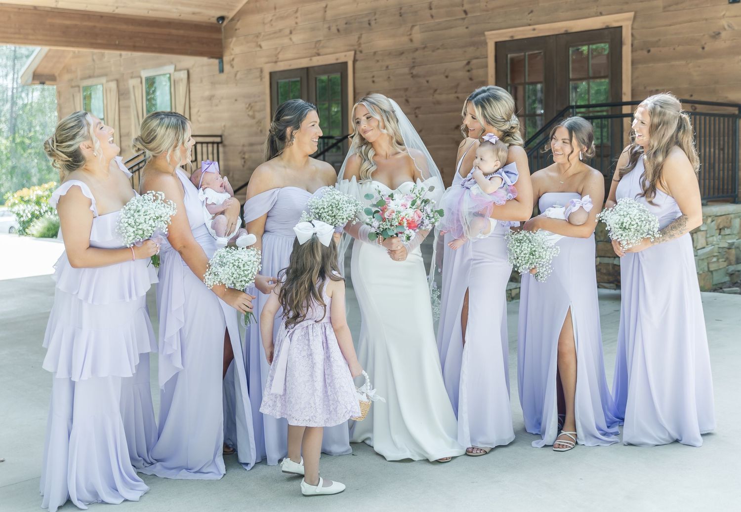 Wedding party wearing light purple dresses gather with white bouquets against wooden lodge exterior.