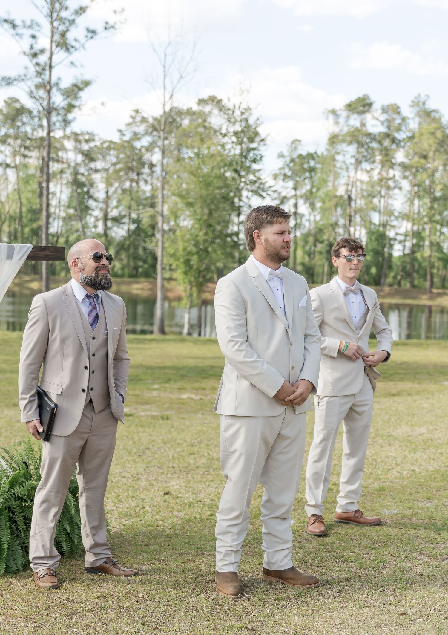 Three groomsmen in coordinating light suits stand during outdoor wedding ceremony near a pond.