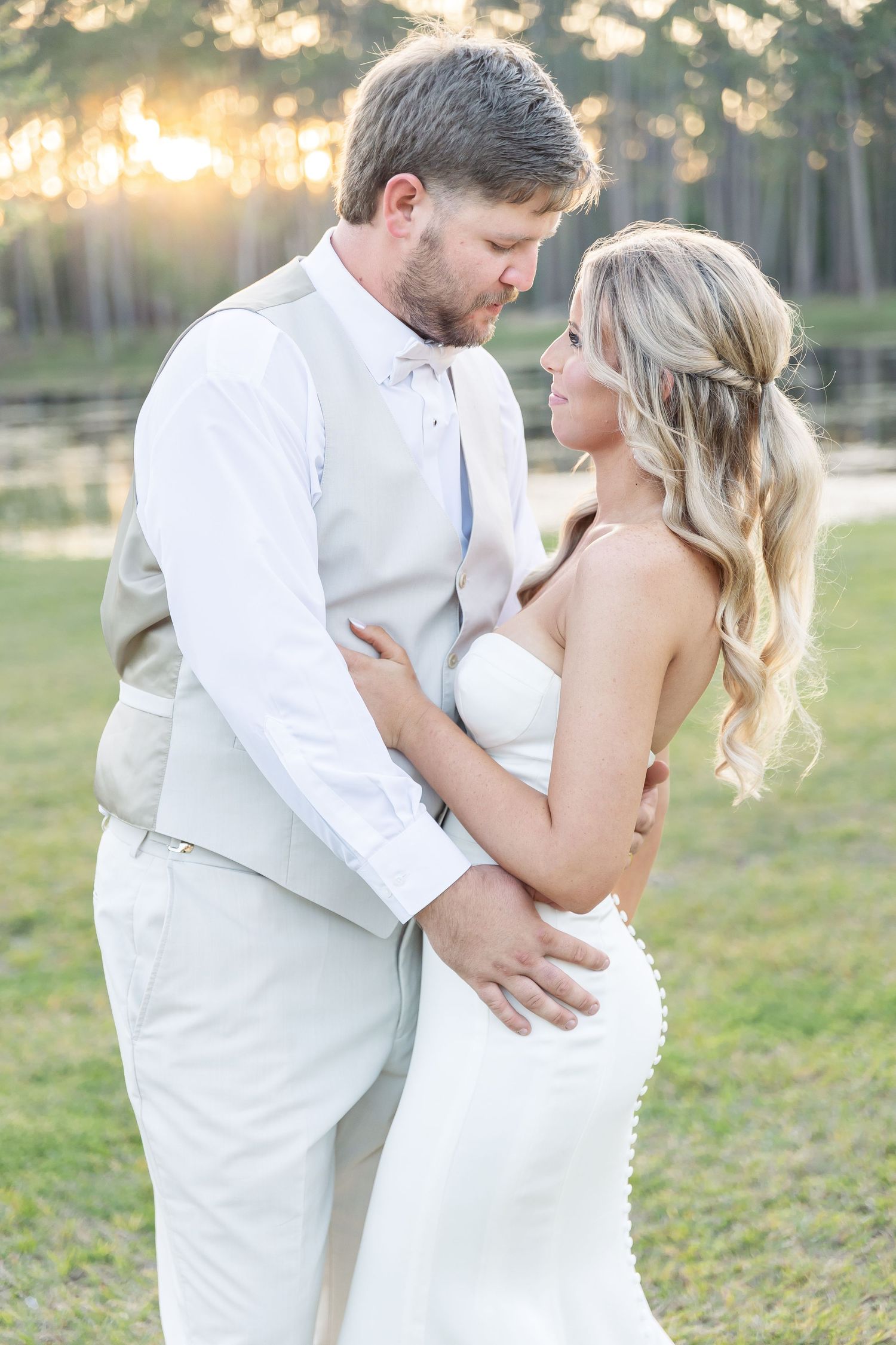 A romantic wedding photo of a couple in white attire embracing at sunset with a lake and trees in the background.