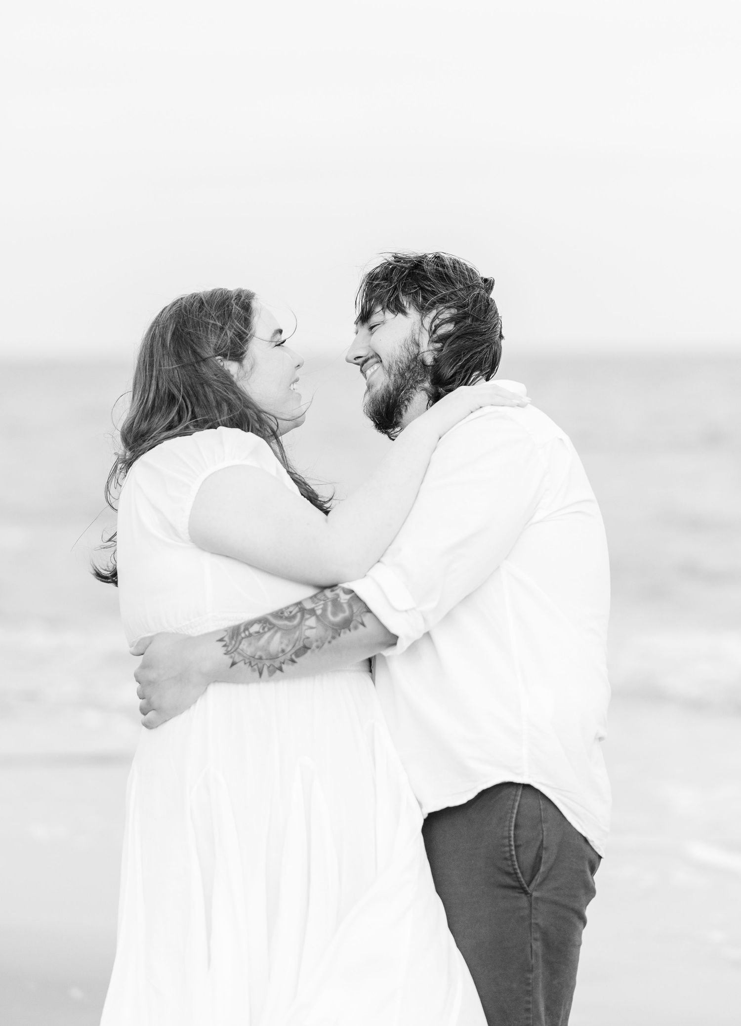 A romantic black and white portrait of a couple embracing at the beach.