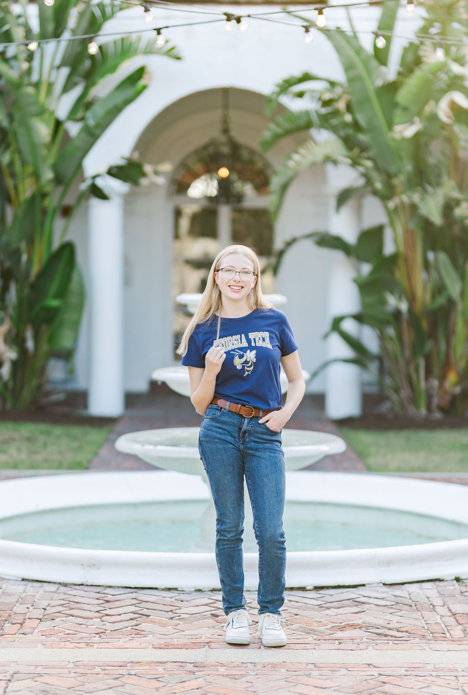 Person stands by fountain and white columns with tropical plants in background.