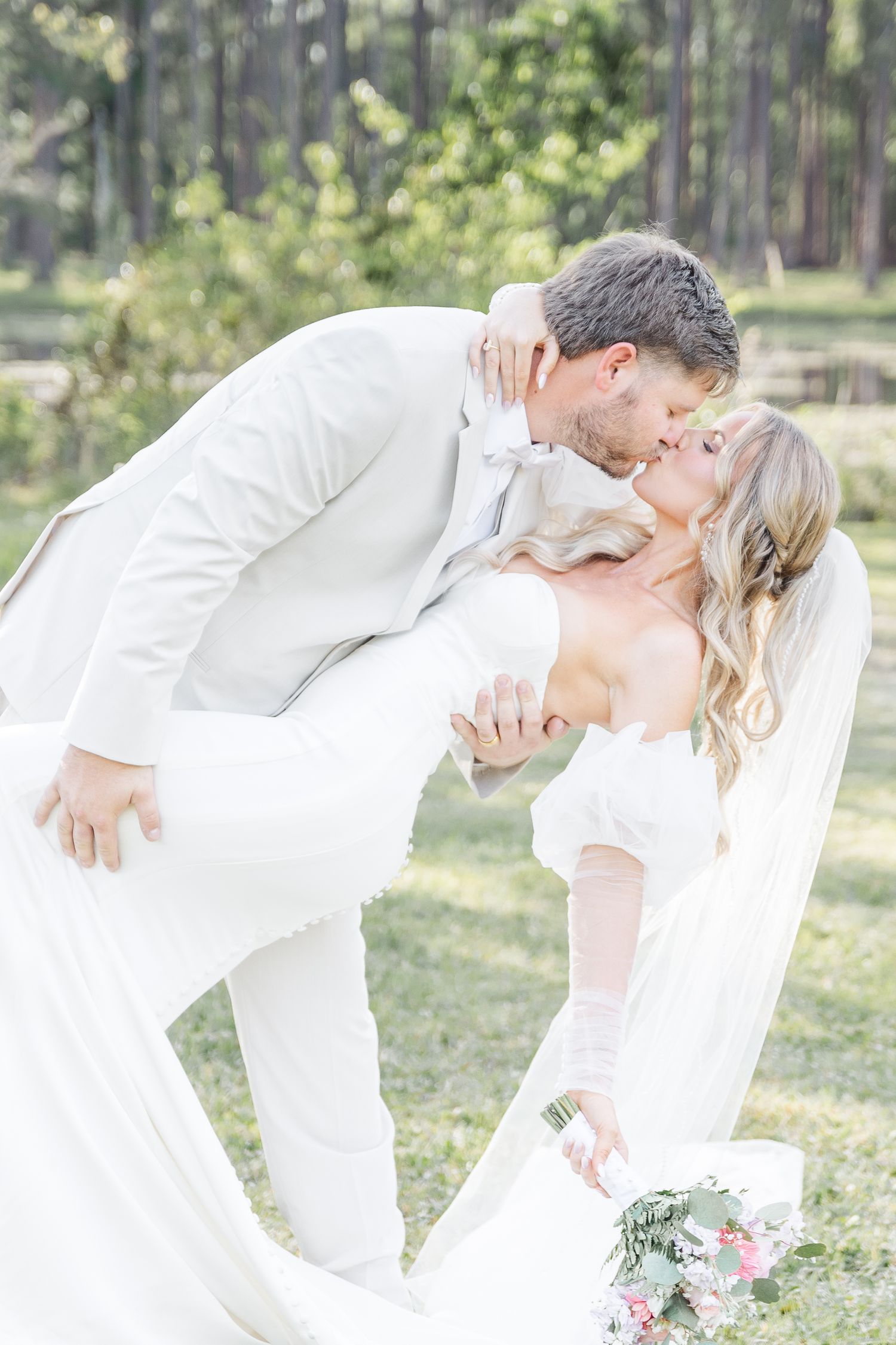 Dramatic dip kiss between bride and groom in matching white outfits during their outdoor wedding photoshoot.