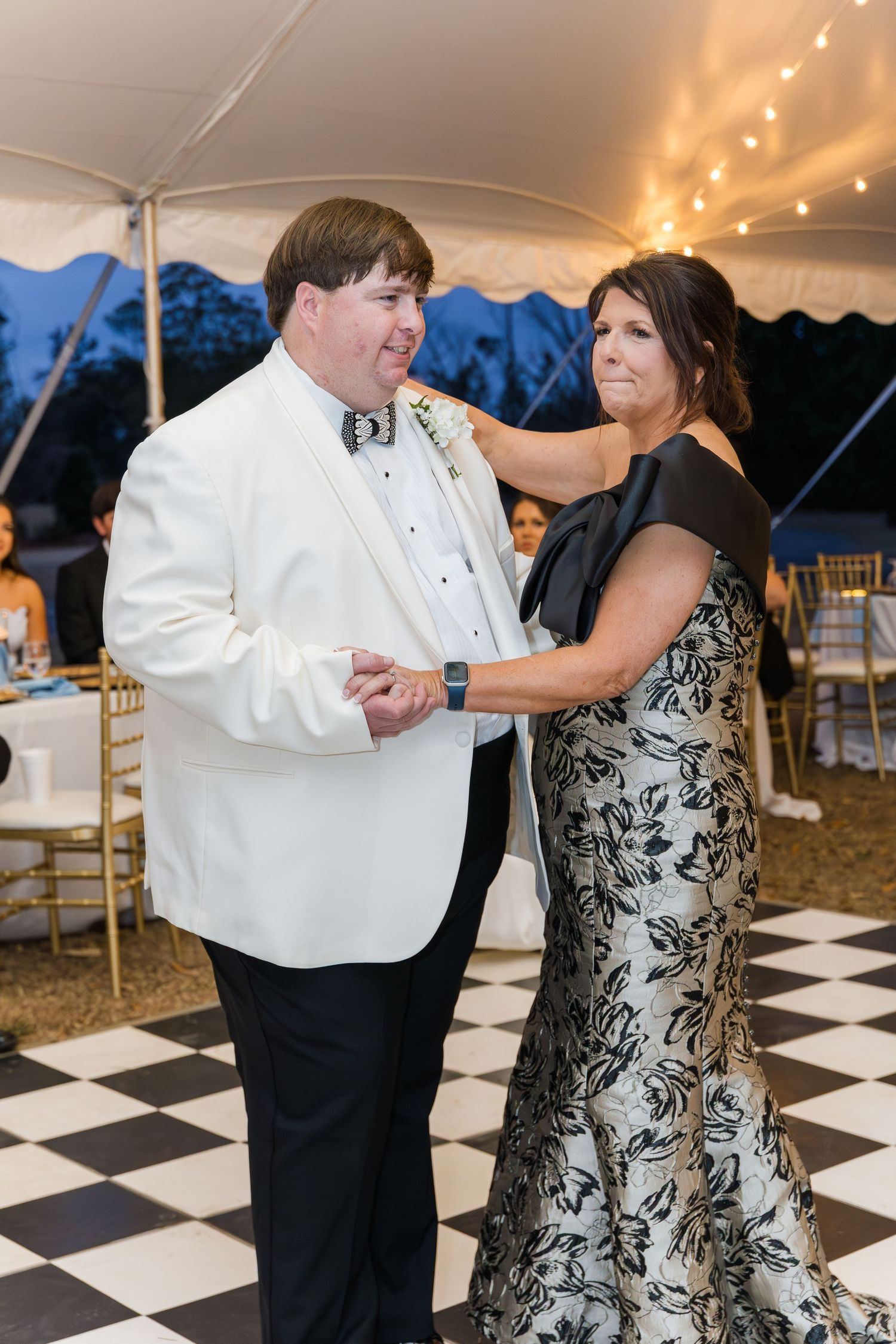 Couple sharing mother-son dance under tented reception with string lights and checkered dance floor.