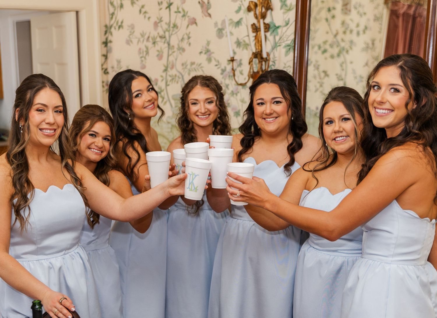 Six bridesmaids in light blue dresses toasting with white cups in a floral wallpapered room.