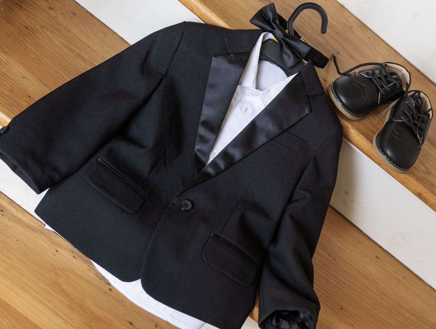 Black tuxedo jacket with white shirt, bow tie on hanger, and small black dress shoes on wooden steps.