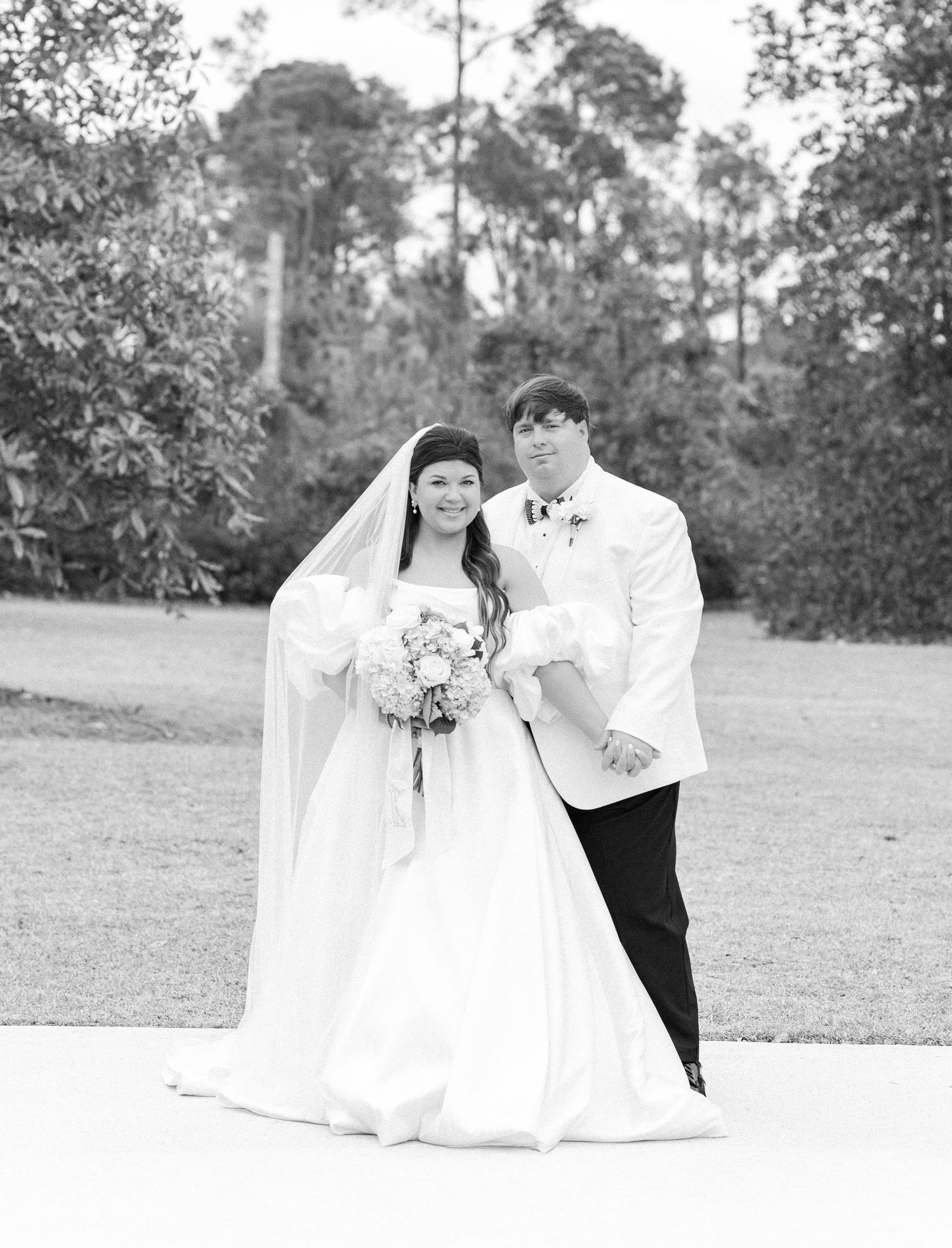 Black and white portrait of bride in ball gown and groom in white jacket on manicured lawn.