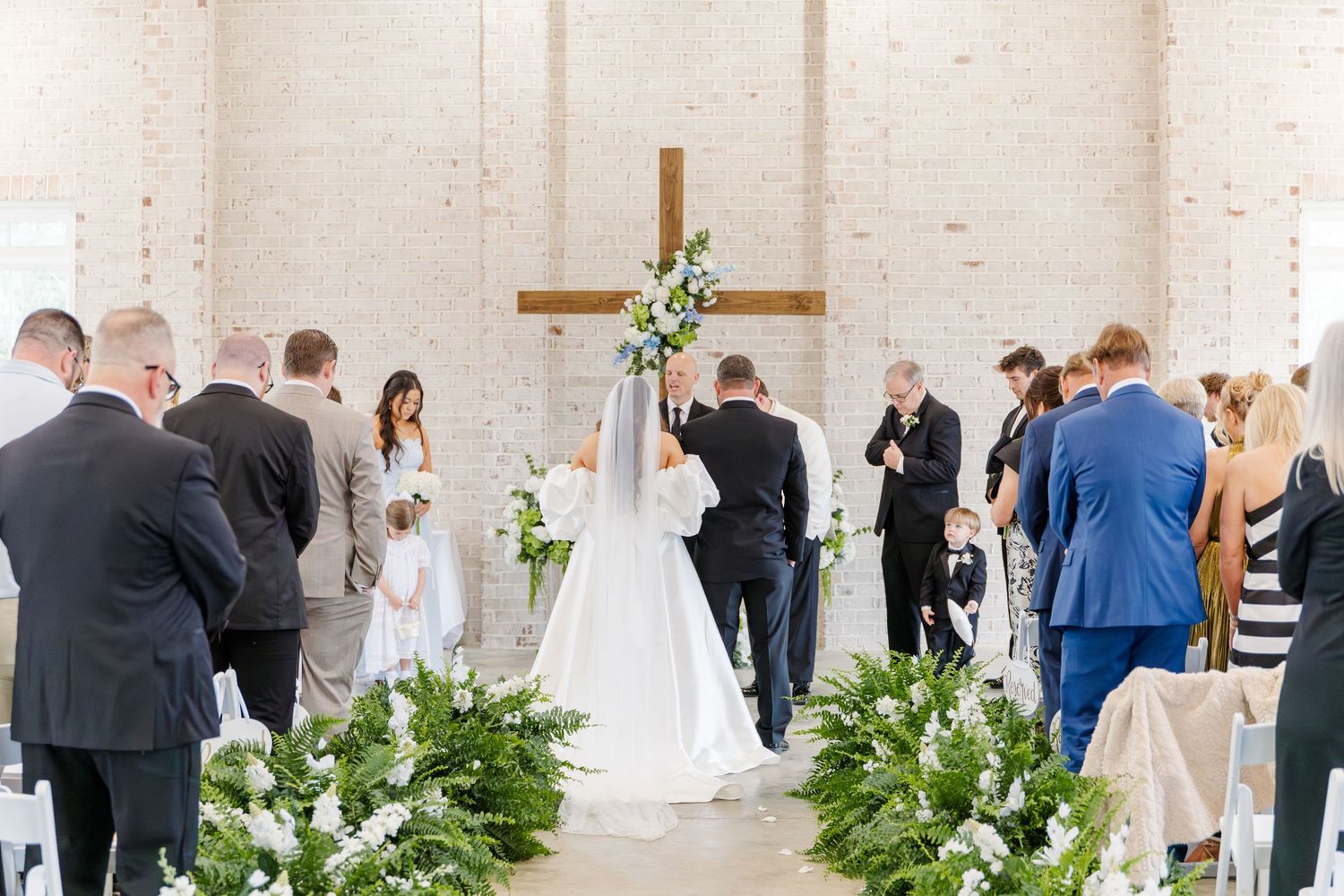 Bride and groom at altar with wooden cross in white brick chapel surrounded by green and white floral arrangements.