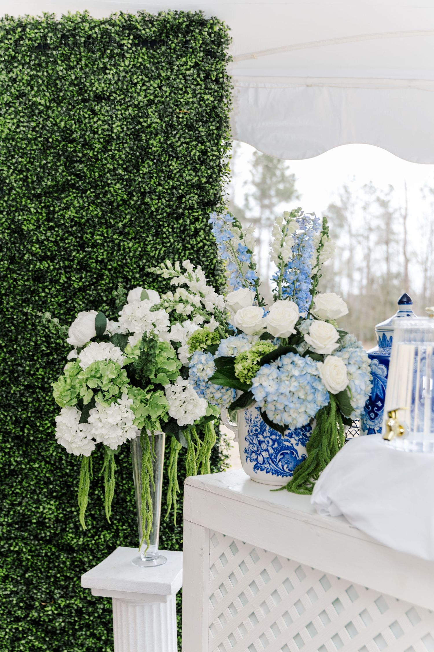 Elegant wedding reception decor with white and blue hydrangea floral arrangements in blue chinoiserie vases against a green boxwood wall.