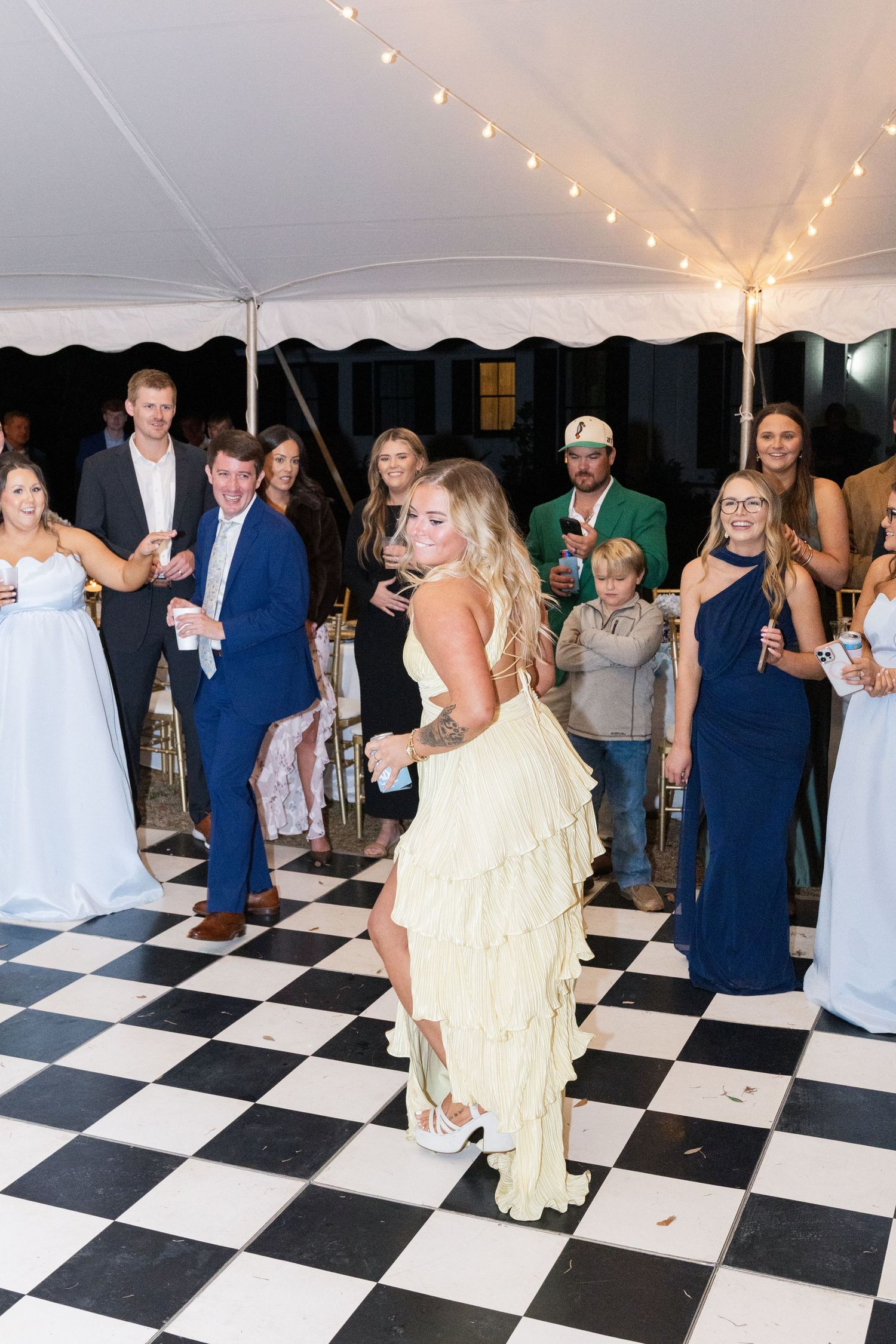 Guest in a yellow ruffled dress dancing on a checkered floor surrounded by wedding reception guests.